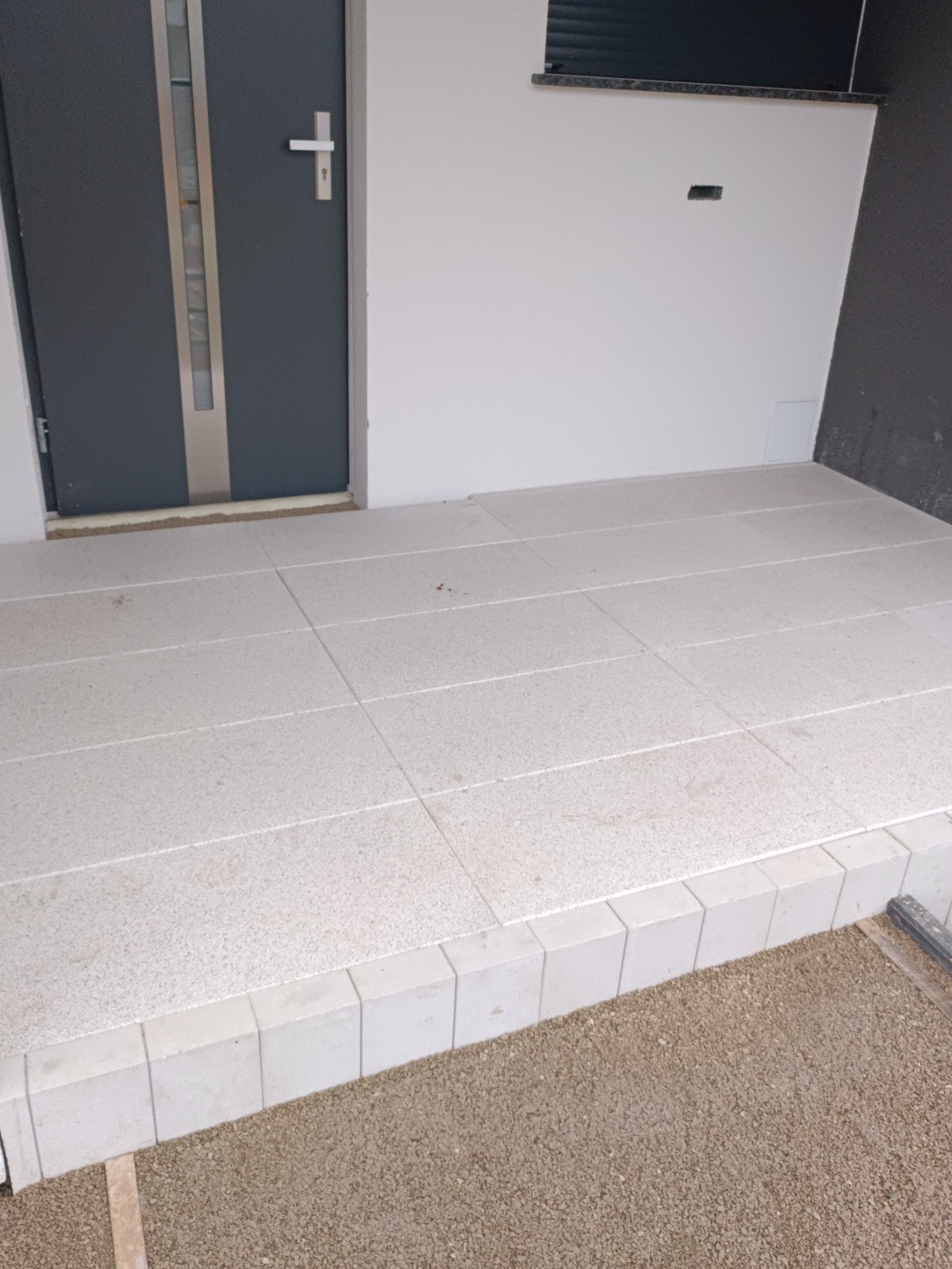 Wykończony taras przed wejściem do domu z jasnych, kwadratowych płytek. Widoczne obrzeże z kostki i fragment podłoża z grysu.