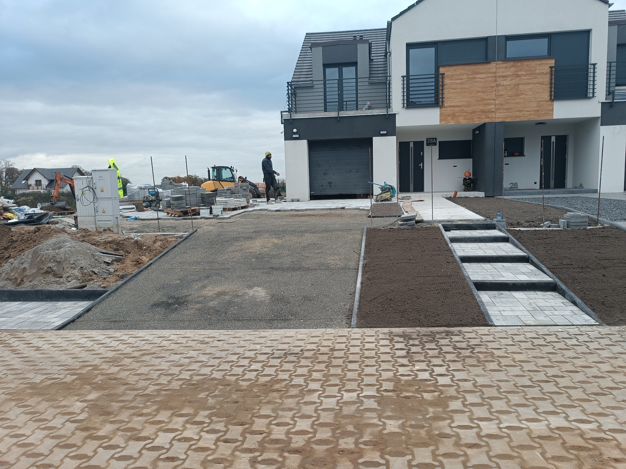 Nowa, częściowo ułożona kostka brukowa przed szeregiem domów. Widoczny sprzęt budowlany i pracownicy. Kostka w różnych kolorach i wzorach. Etap prac w pochmurny dzień.