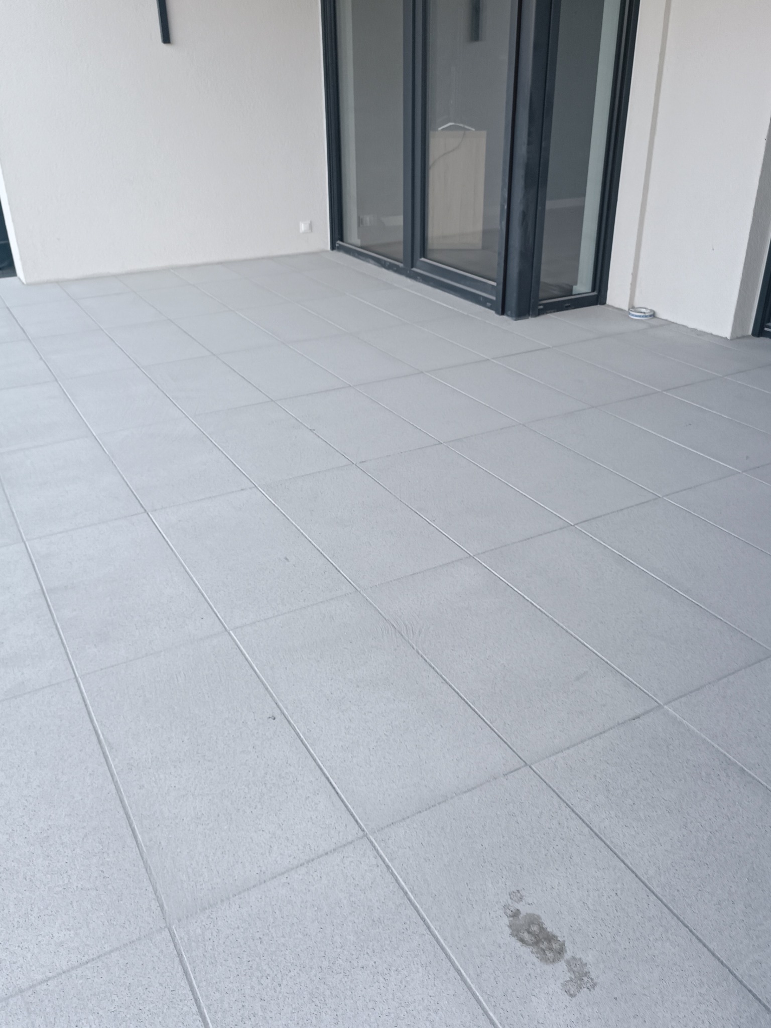 Jasnoszara, kwadratowa płytka na balkonie z widocznym śladem zabrudzenia w prawym dolnym rogu. W tle biała ściana i przesuwne drzwi balkonowe z czarną ramą.