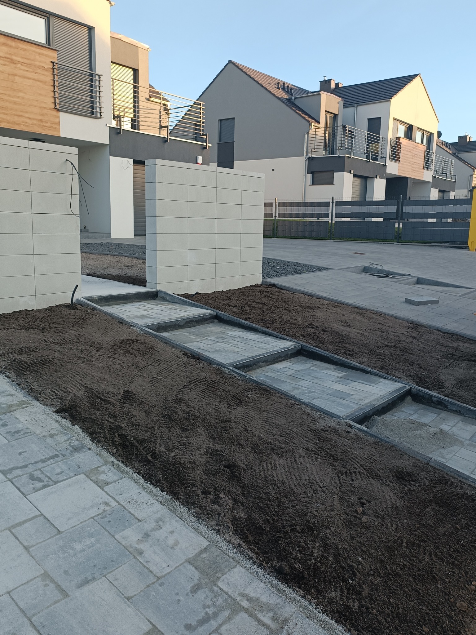 Nowoczesna posesja w trakcie realizacji: szara kostka brukowa tworzy ścieżkę przez rabaty z ziemią, na tle segmentów mieszkalnych z balkonami.