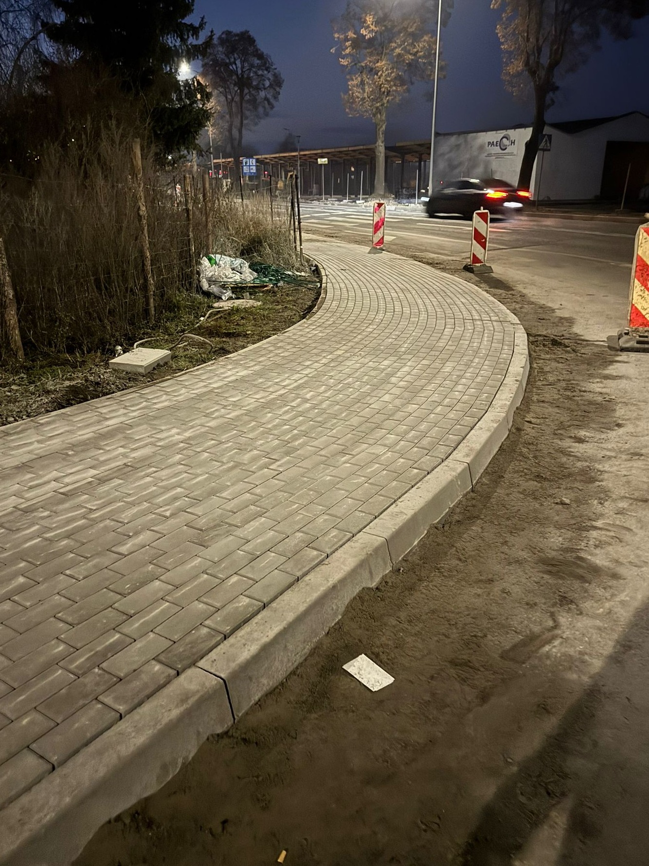 Zakrzywiony chodnik z szarej kostki brukowej, obrzeża betonowe, w tle ulica i budynki. Widoczne słupki ostrzegawcze i pozostałości po pracach ziemnych.