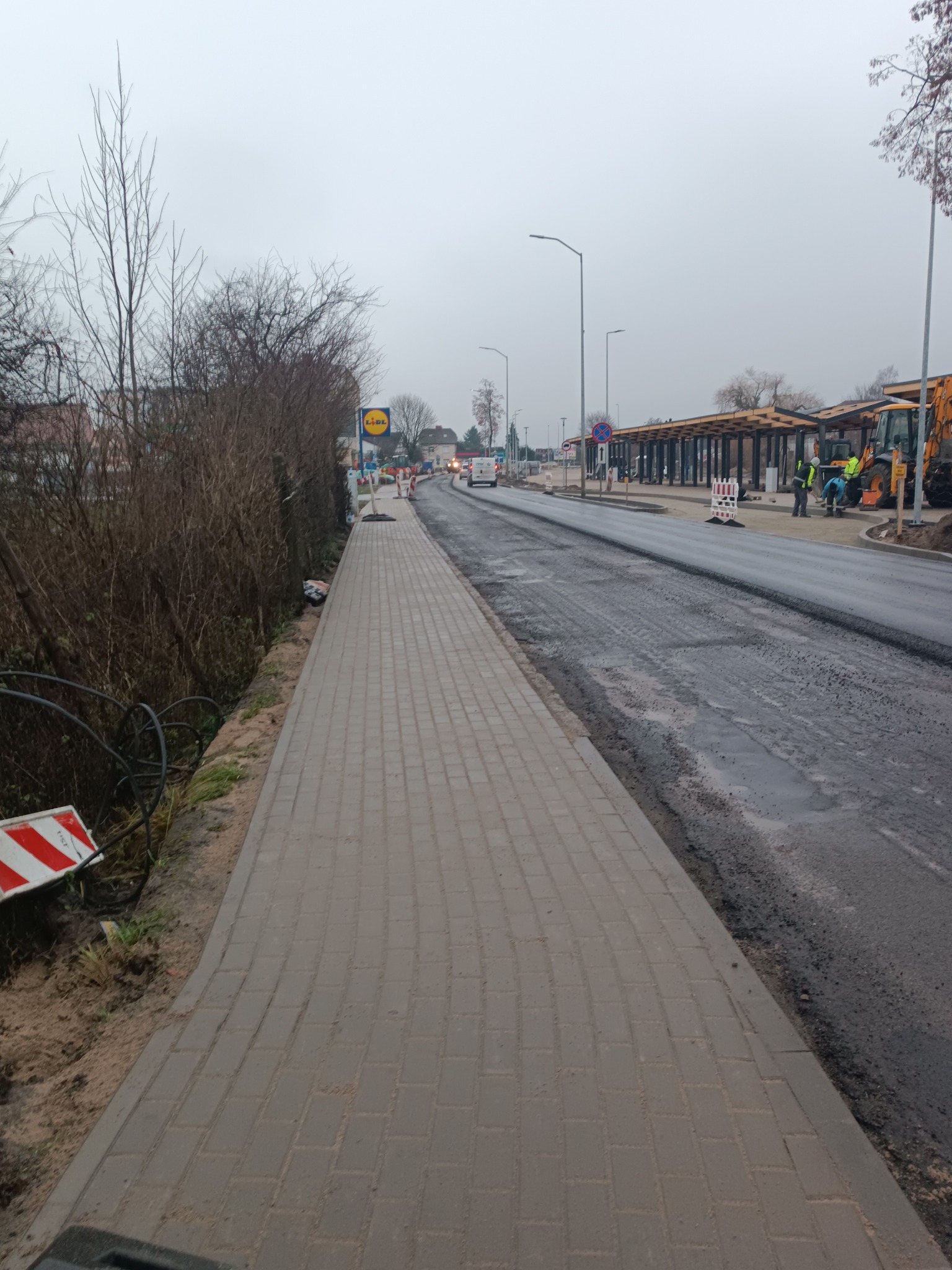 Ułożona kostka brukowa chodnika wzdłuż ulicy z budowaną zatoką autobusową i pracującymi maszynami budowlanymi w tle, pochmurny dzień.