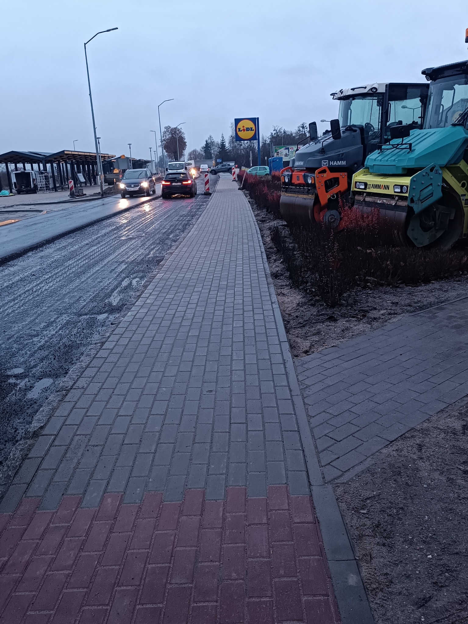 Ułożona kostka brukowa w szarym i czerwonym kolorze, obok walce drogowe i świeżo wylany asfalt. W tle widoczny znak Lidl i przejeżdżające samochody.
