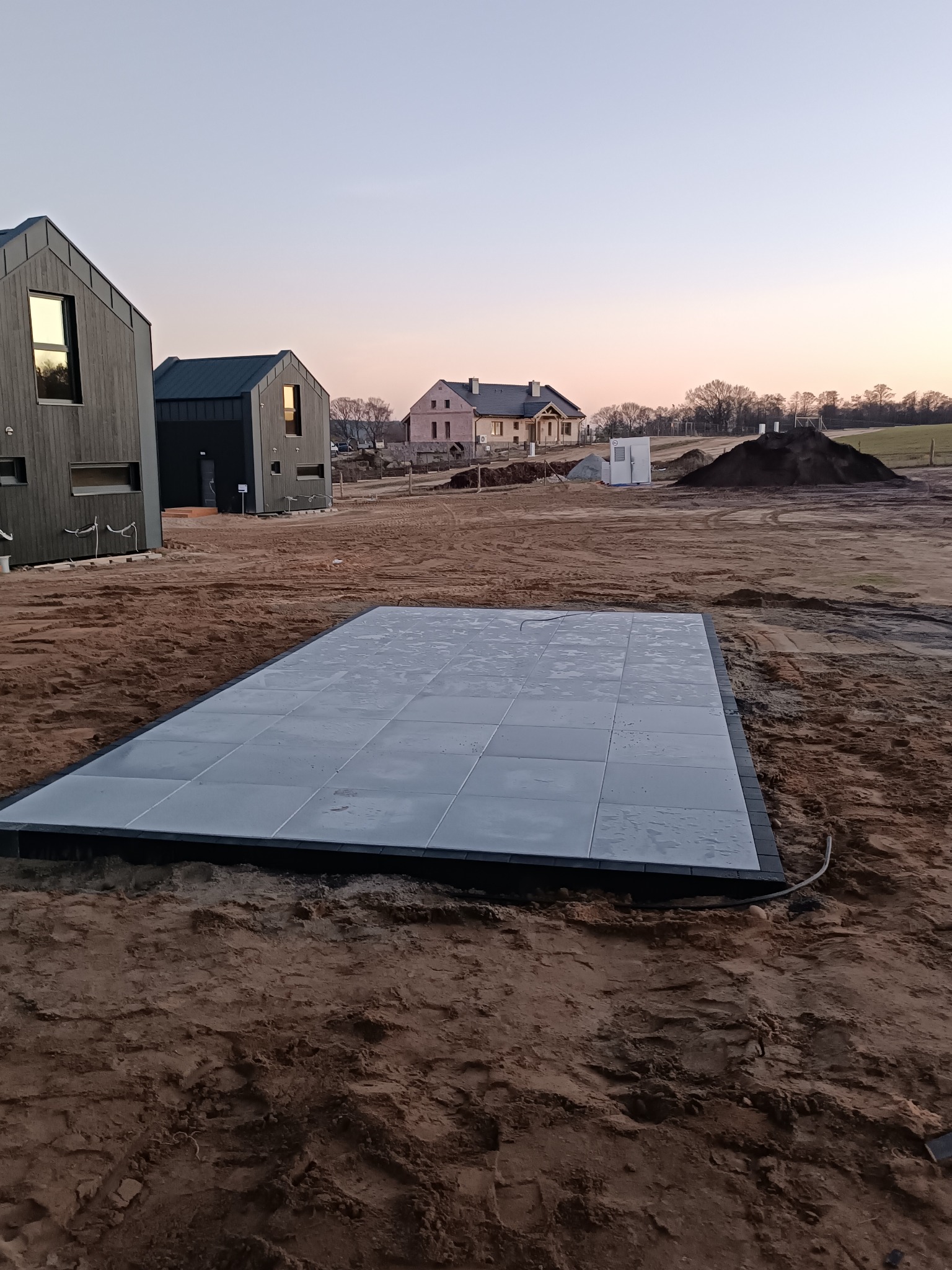 Ułożona, prostokątna nawierzchnia z szarych płyt w Gorzowie Wielkopolskim. W tle domy w budowie i zachodzące słońce. Widok z poziomu gruntu.