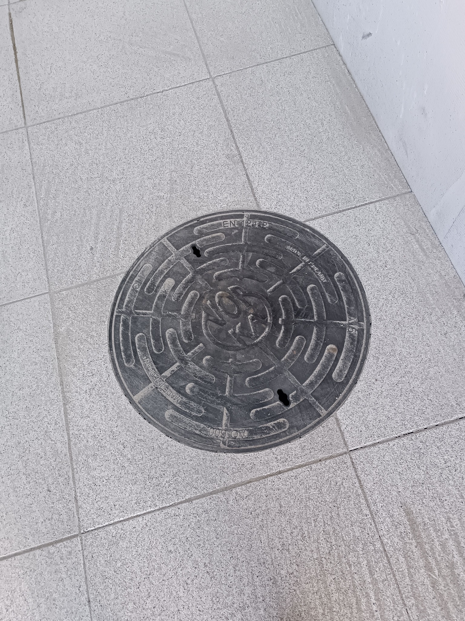 Szara, okrągła studzienka kanalizacyjna z napisem 'Made in Poland' wkomponowana w jasnoszarą, kwadratową kostkę brukową przy białej ścianie.