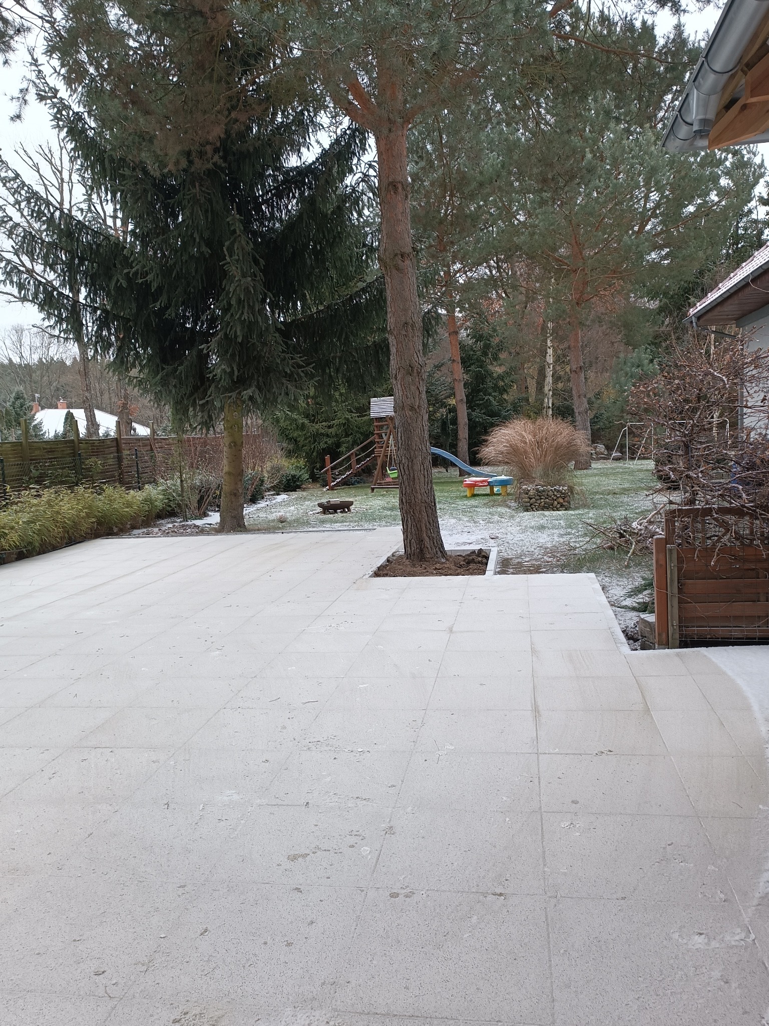 Jasna, nowa nawierzchnia z płyt na zewnątrz domu, z drzewem rosnącym w wyciętej przestrzeni. W tle ogród z placem zabaw i elementami drewnianego ogrodzenia.