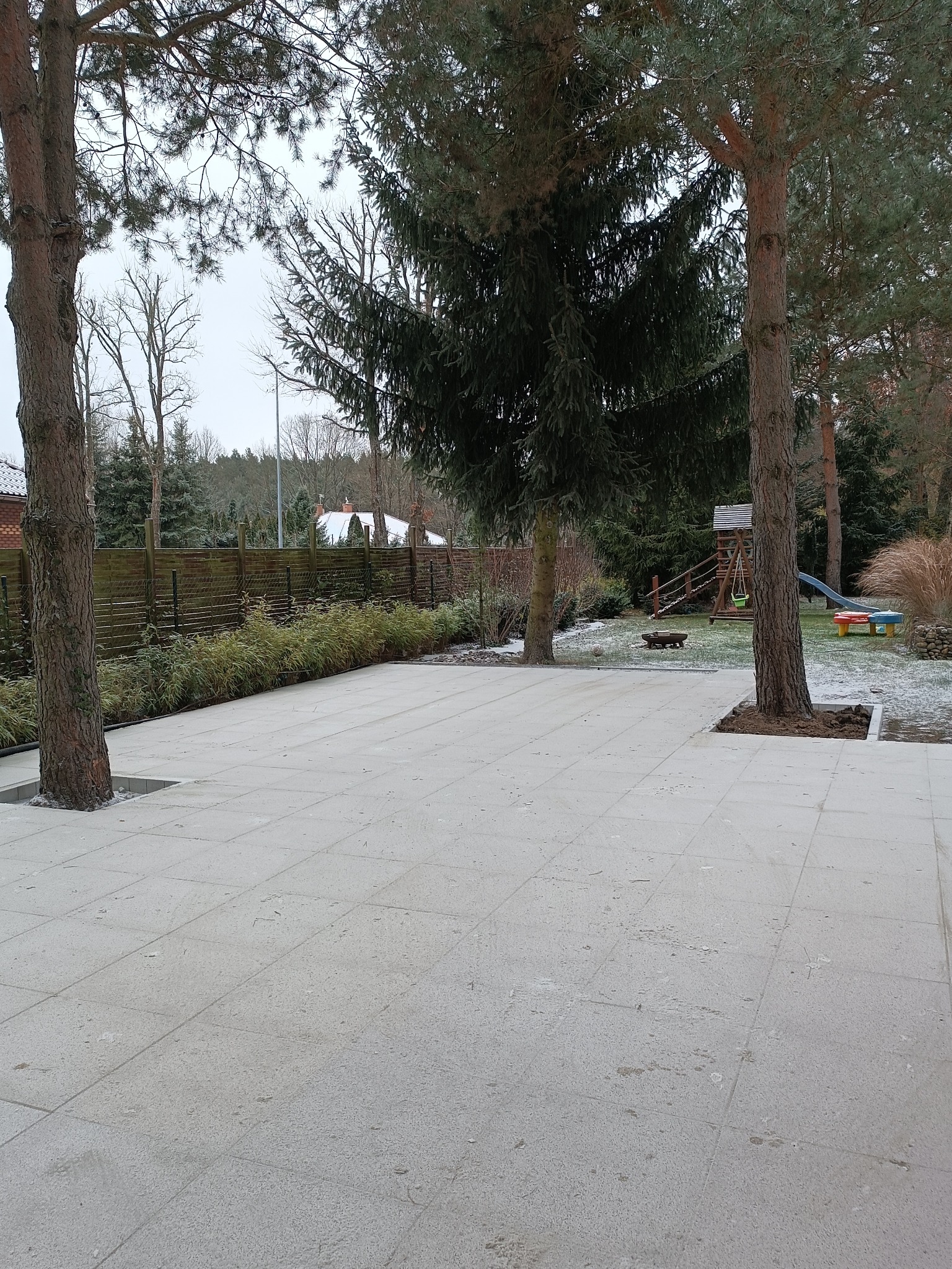Jasnoszara kostka brukowa ułożona wokół drzew na posesji. W tle widoczny drewniany płot i plac zabaw.