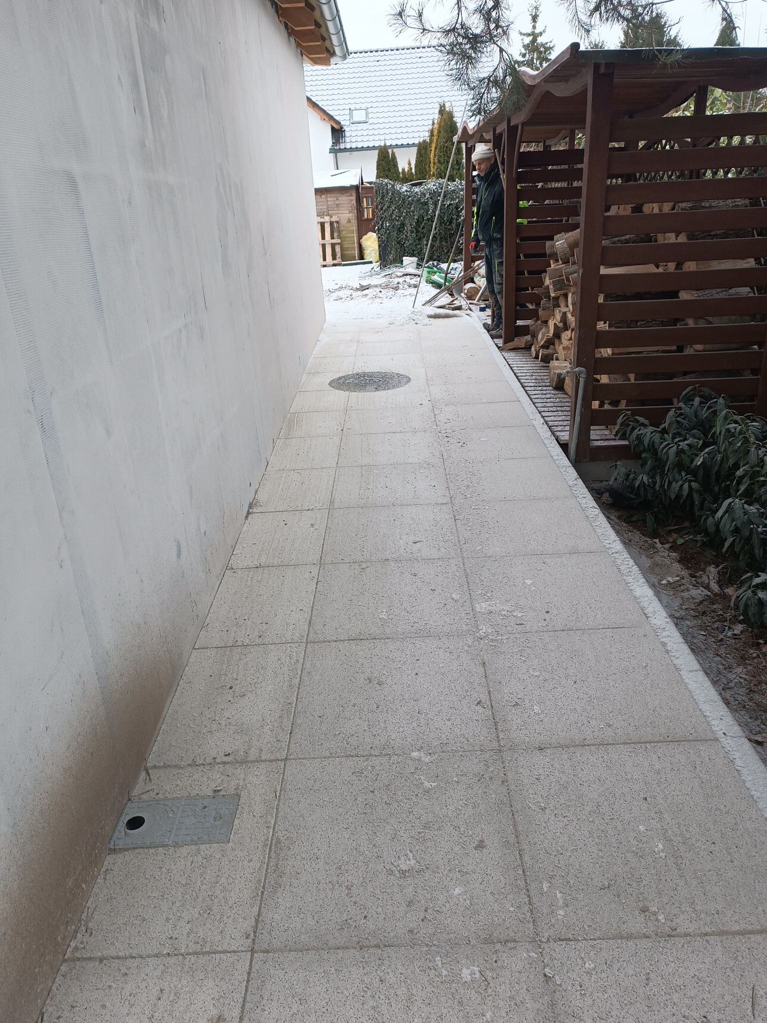 Ułożona, jasnoszara kostka brukowa tworząca ścieżkę wzdłuż ściany budynku. Widoczny właz kanalizacyjny i drewniana wiata na drewno. Praca w toku, widoczny pracownik.