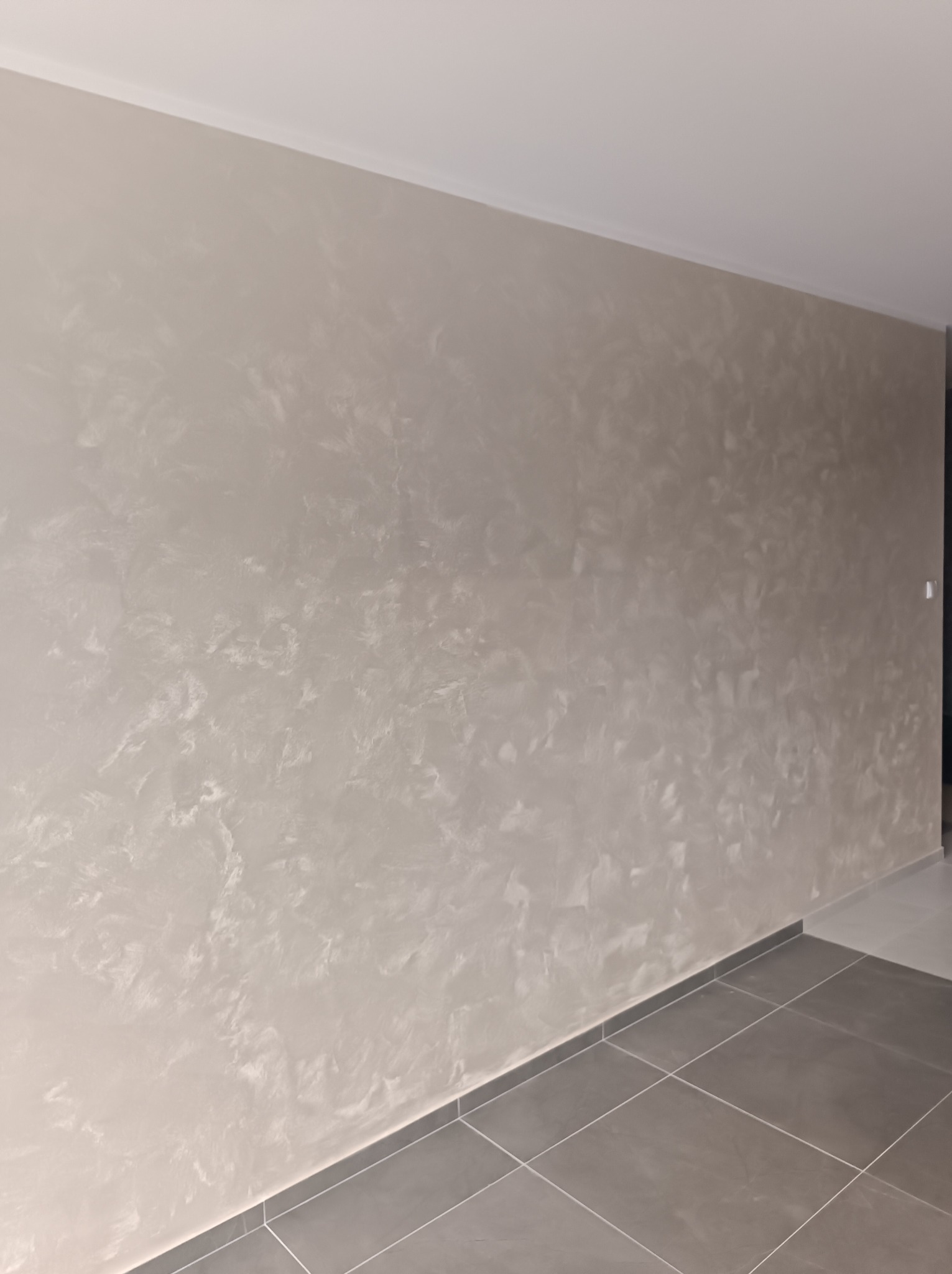 Ściana z dekoracyjną, beżową strukturą przypominającą marmur, wykończona listwą przysufitową. Szare płytki podłogowe z fugą. Minimalistyczny design wnętrza.