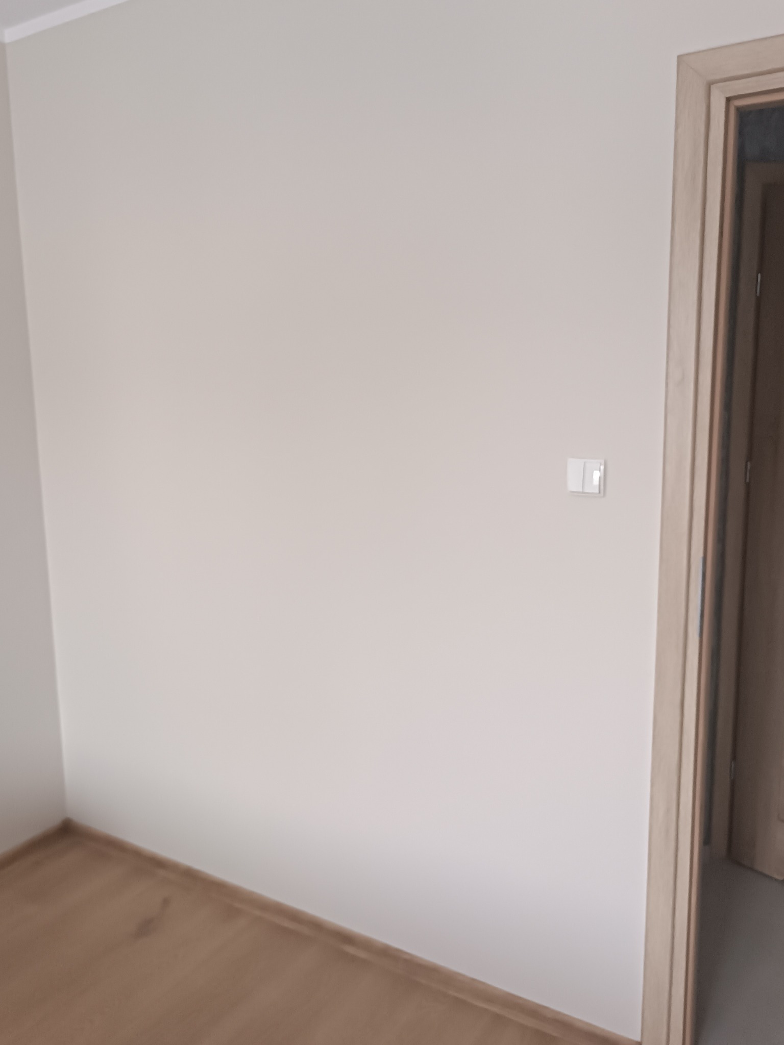 Jasny pokój po malowaniu: gładkie, beżowe ściany, drewniana podłoga i rama drzwi. Widoczny włącznik światła. Minimalistyczne wykończenie wnętrza.