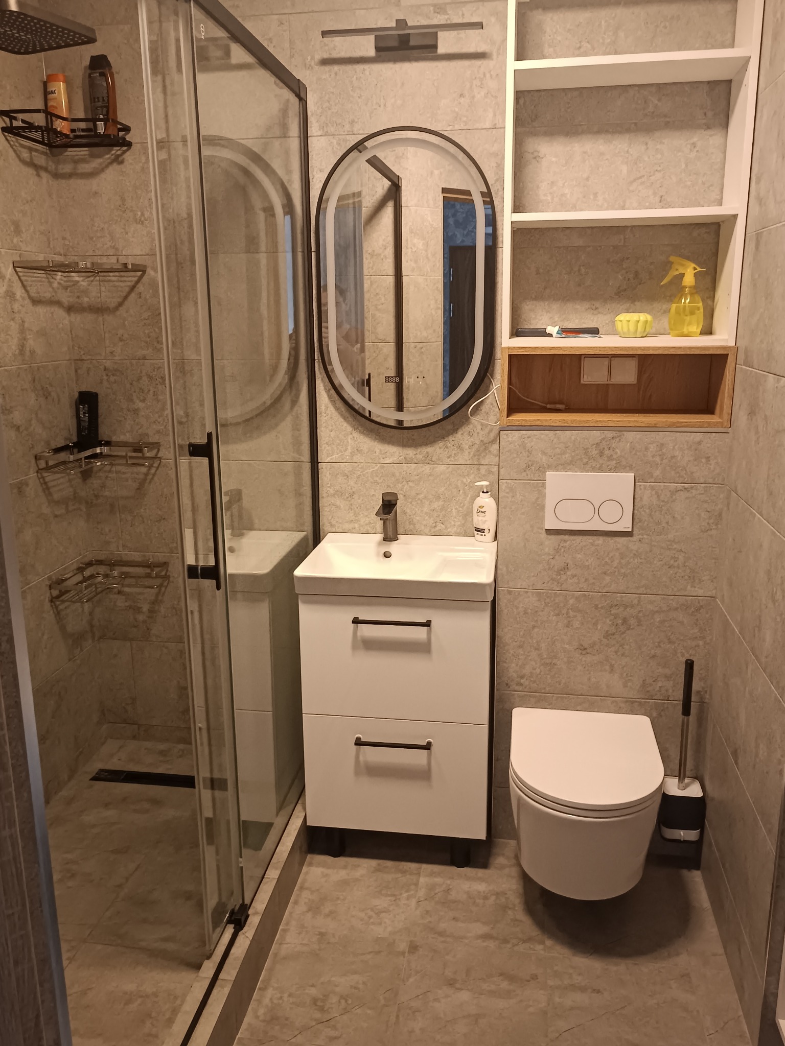 Nowoczesna łazienka z kabiną prysznicową, umywalką z szafką, owalnym lustrem z podświetleniem i podwieszaną toaletą. Szare płytki imitujące kamień na ścianach i podłodze.