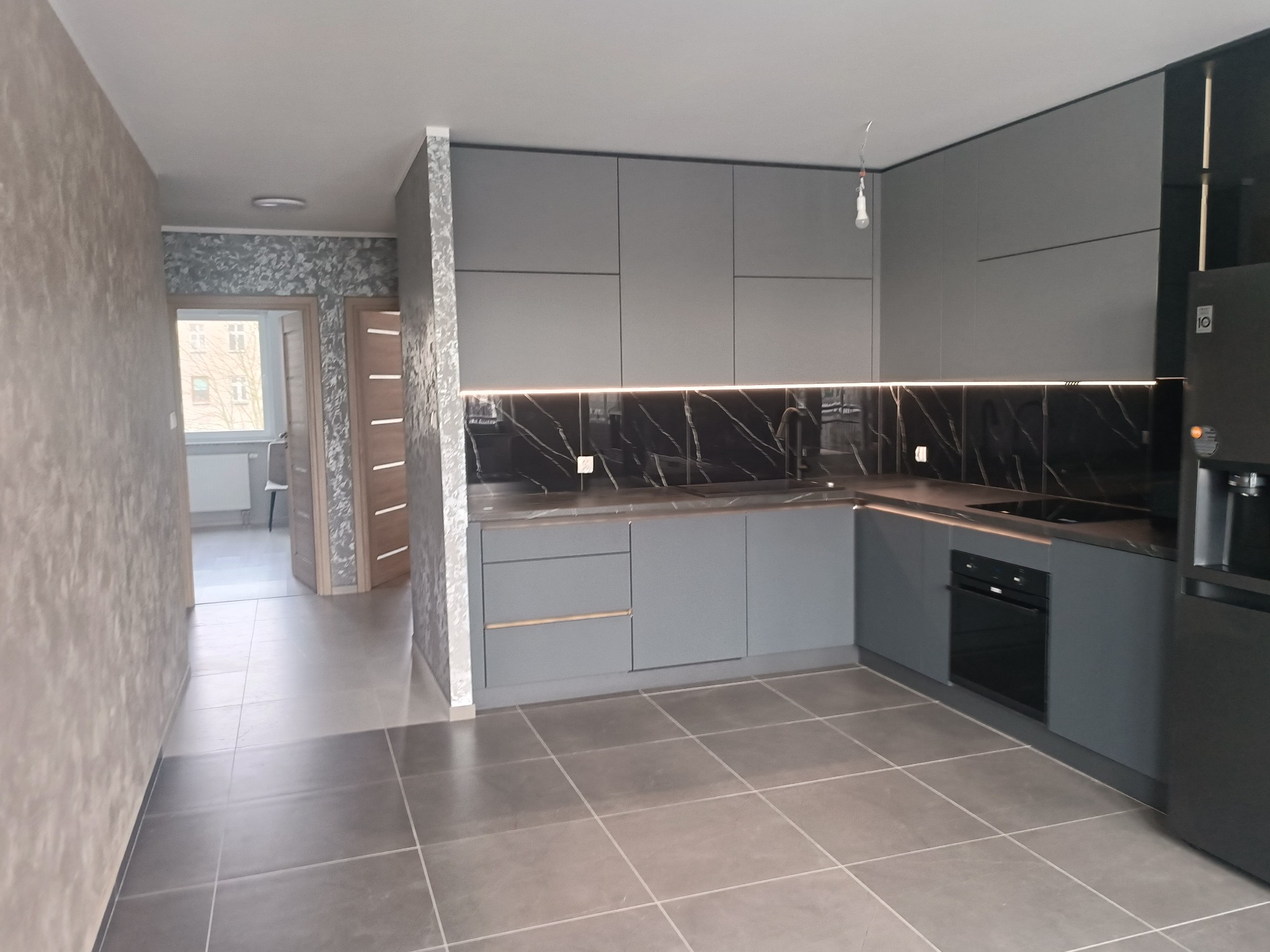 Szara kuchnia z marmurowym blatem i podświetleniem LED, widok na korytarz z dekoracyjną tapetą. Nowoczesne wykończenie wnętrz.