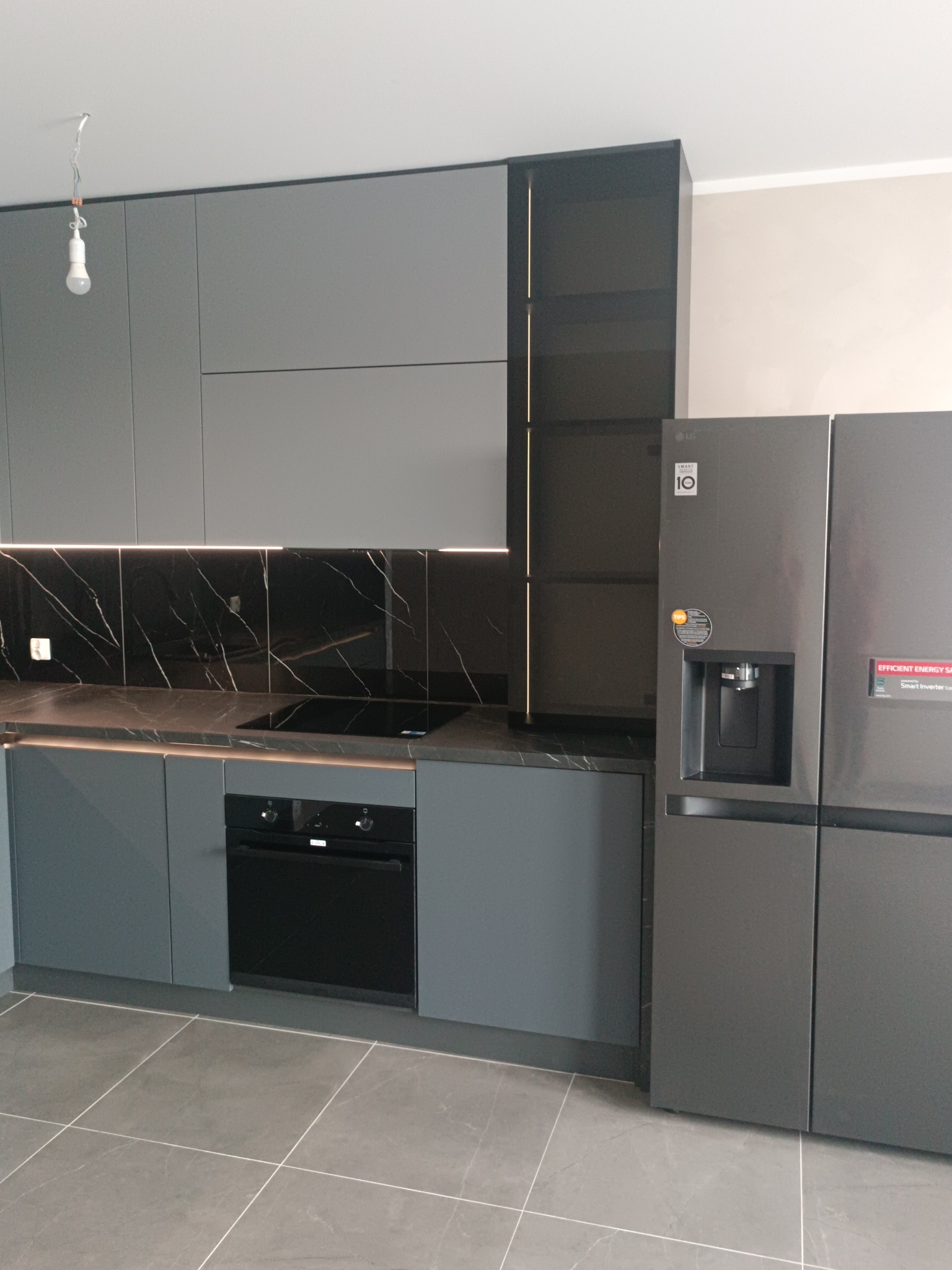 Nowoczesna kuchnia w odcieniach szarości z marmurowym blatem i podświetleniem LED. Widoczna zabudowa kuchenna, piekarnik i lodówka side-by-side.