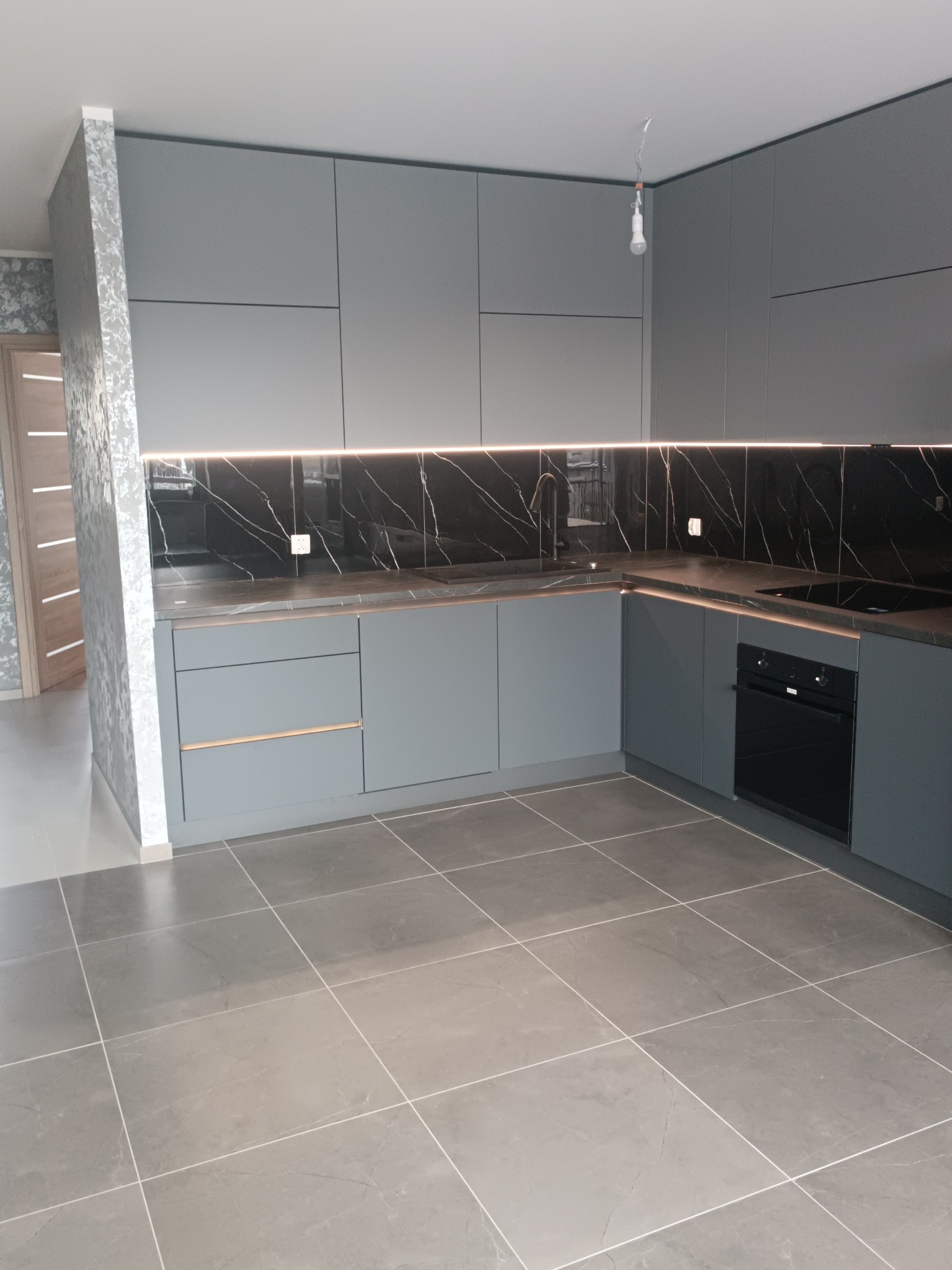 Nowoczesna kuchnia w szarościach z czarnym marmurem na ścianie, podświetleniem LED i szarą podłogą z płytek. Minimalistyczny design, widok na fragment drzwi.