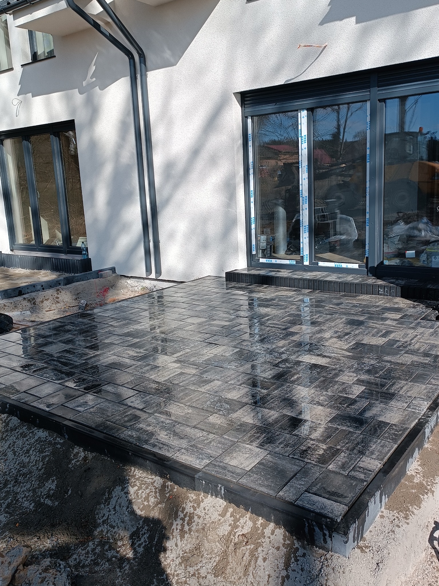 Nowy taras z kostki brukowej w odcieniach szarości, przylegający do nowoczesnego domu z dużymi oknami. Widoczne detale wykończenia i świeżo ułożona powierzchnia.