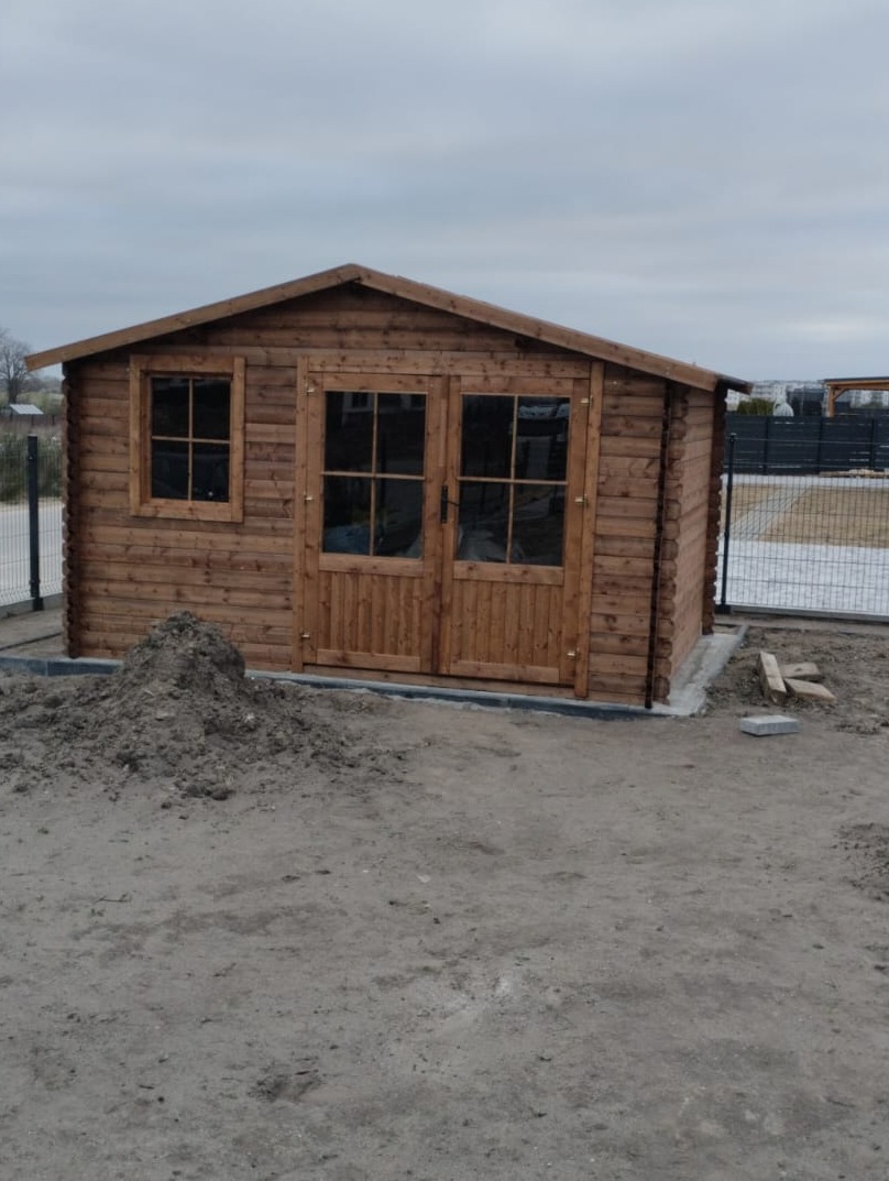 Drewniany domek ogrodowy z dwuskrzydłowymi drzwiami i oknem, otoczony ziemią i fragmentem ogrodzenia. Widoczne detale konstrukcji i naturalne wykończenie drewna.