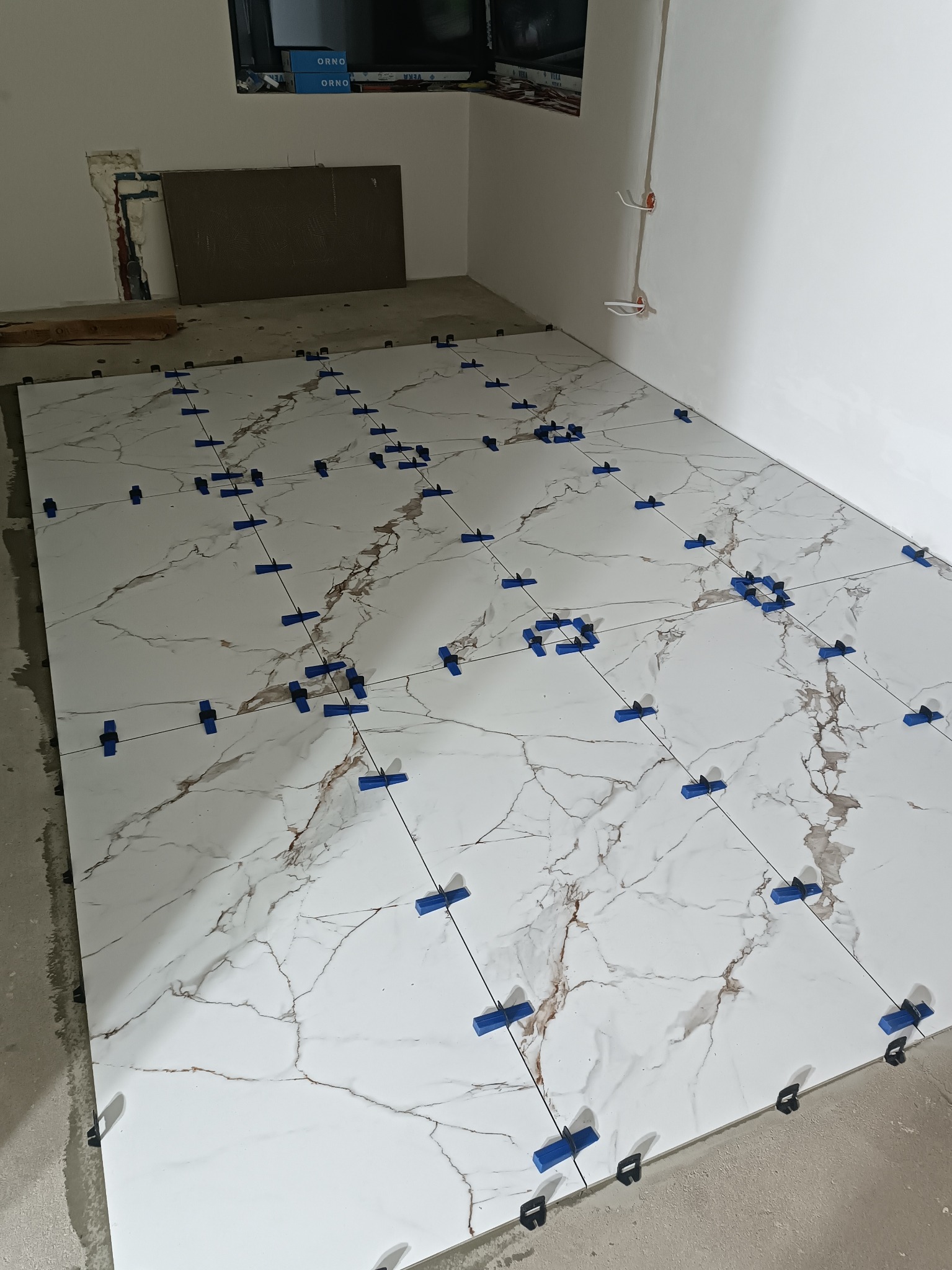 Białe płytki imitujące marmur, ułożone na podłodze z użyciem niebieskich klipsów poziomujących. Widoczne kable elektryczne w ścianie i surowy stan wykończenia.