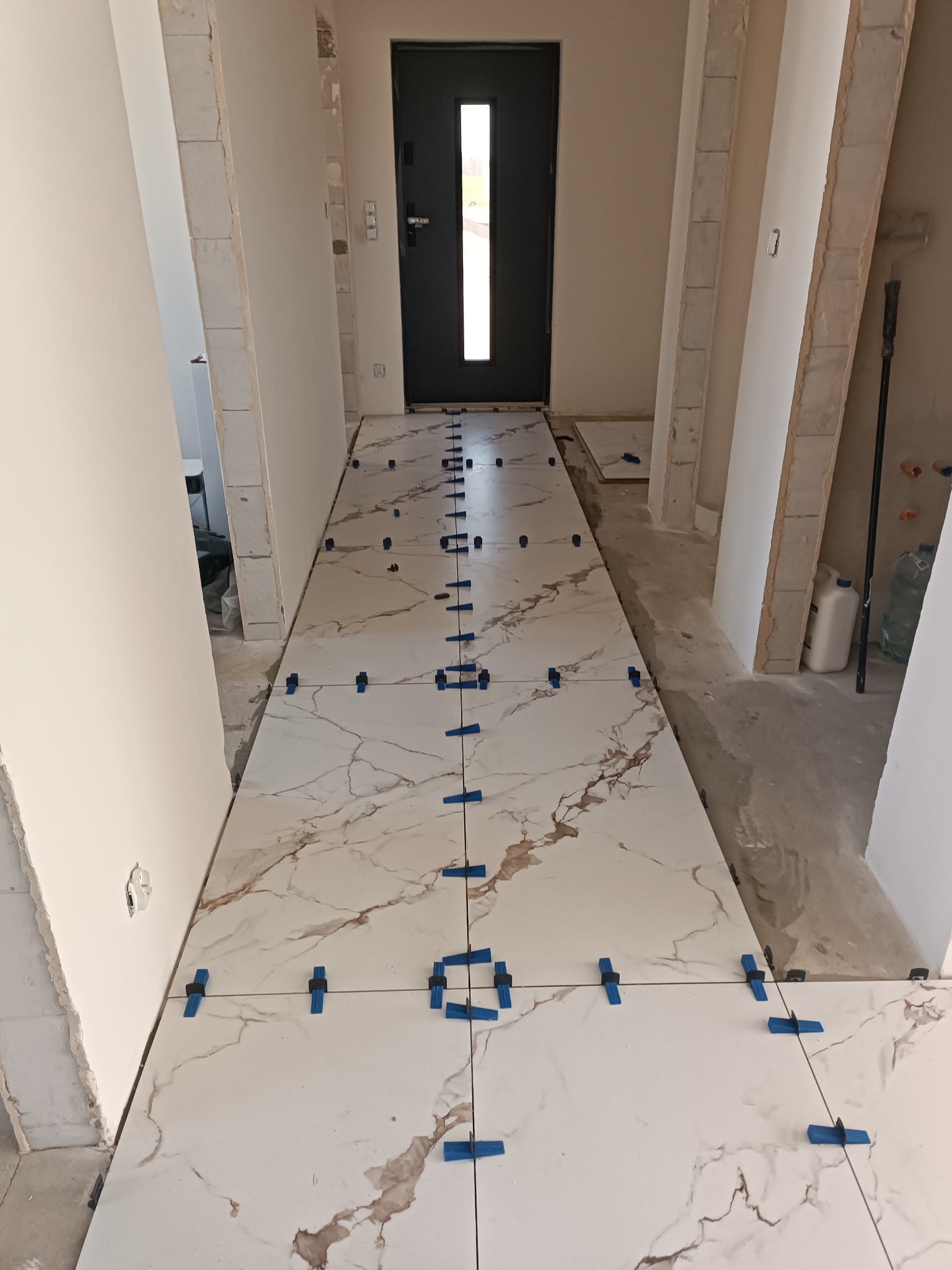Białe płytki imitujące marmur, ułożone w korytarzu, z niebieskimi klipsami poziomującymi. Widoczny fragment drzwi wejściowych i surowy stan ścian.