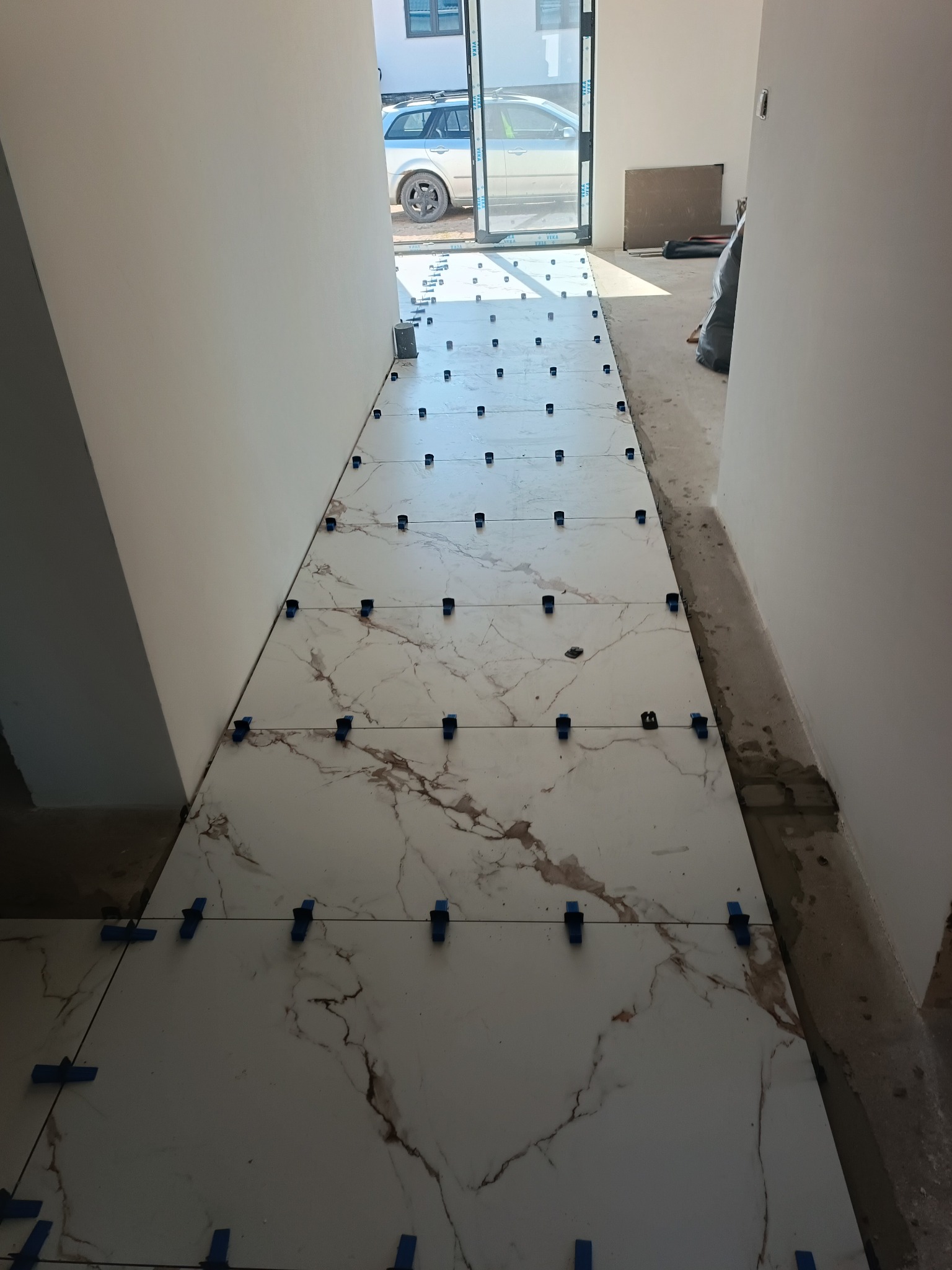 Białe płytki imitujące marmur, ułożone w korytarzu, z niebieskimi klipsami poziomującymi. Widok na zewnątrz przez otwarte drzwi.