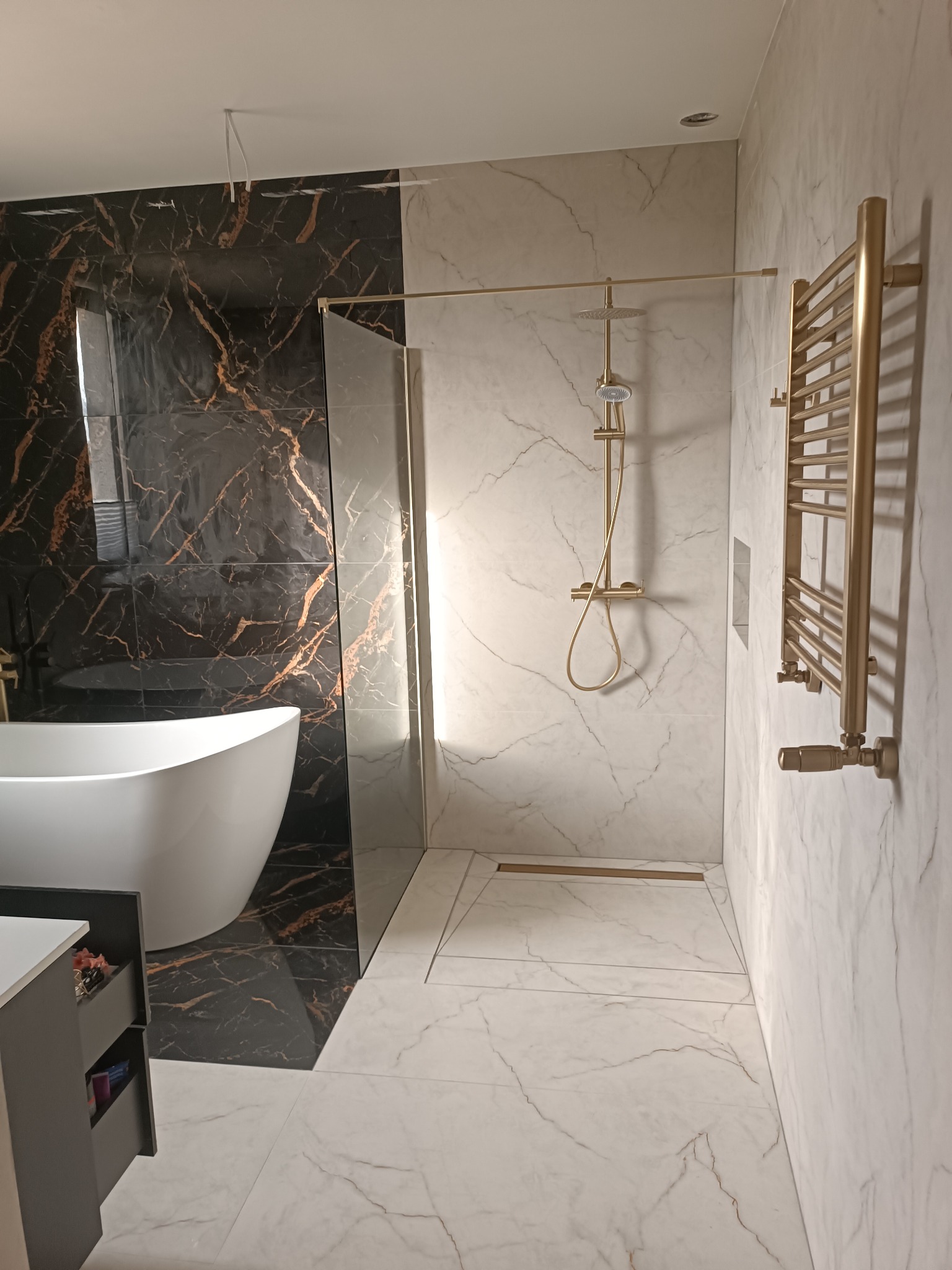 Elegancka łazienka z wanną wolnostojącą, kabiną prysznicową typu walk-in i grzejnikiem drabinkowym w kolorze złotym. Marmurowe płytki na ścianach i podłodze.