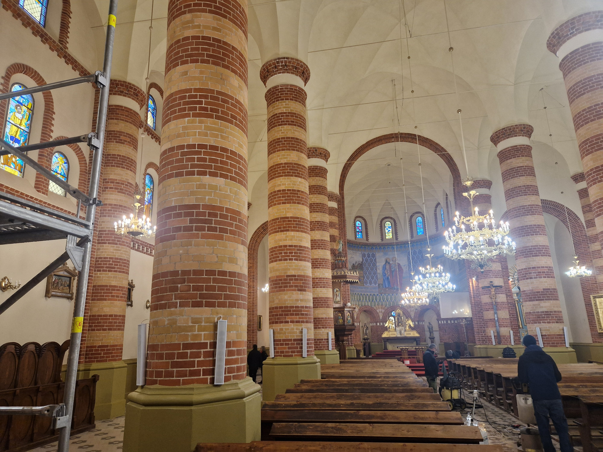 Wnętrze kościoła z ceglanymi kolumnami i drewnianymi ławkami, widoczne rusztowania i prace renowacyjne w toku. Charakterystyczne oświetlenie i witraże.