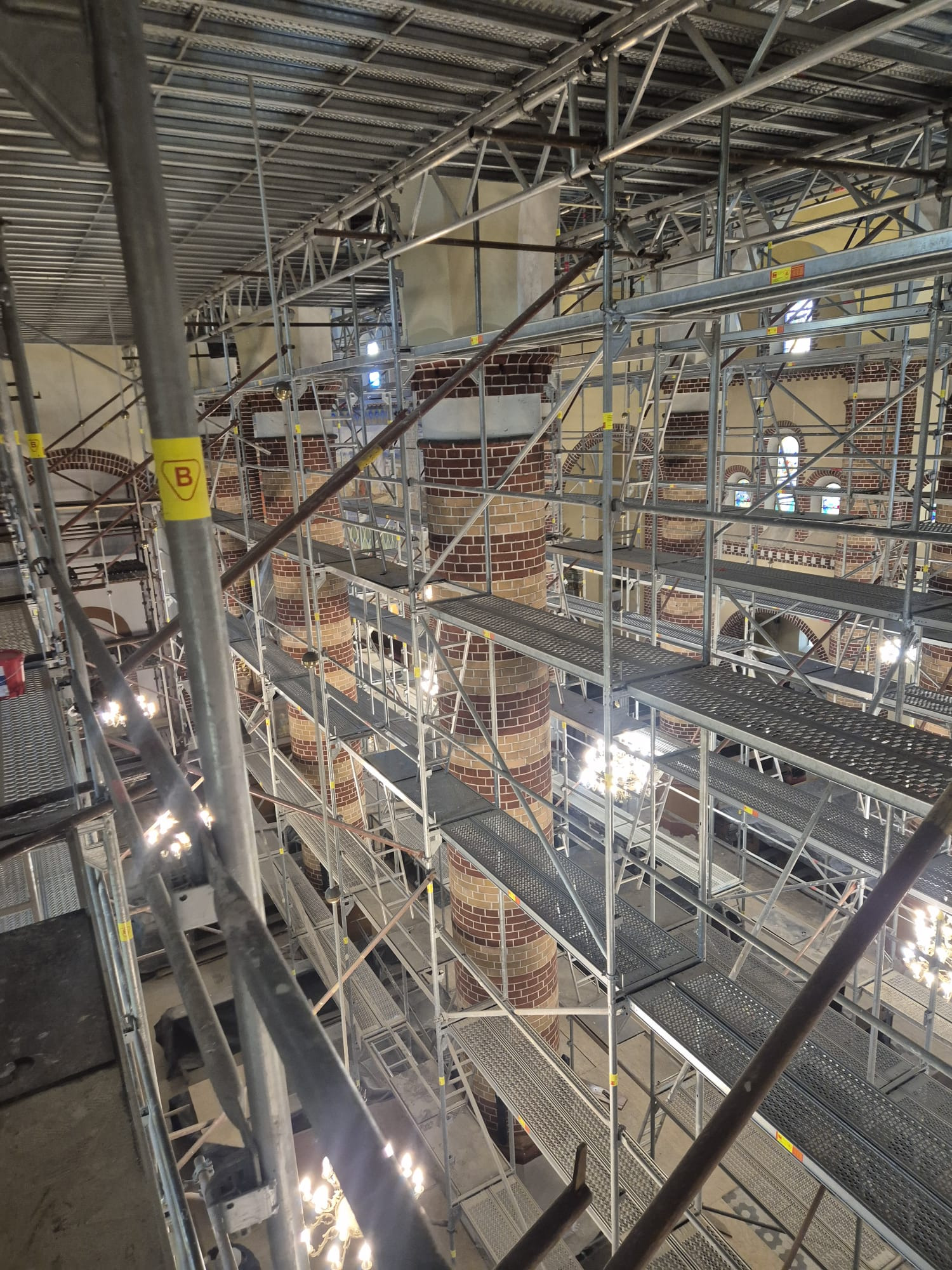 Wnętrze remontowanej kamienicy z kolumnami obłożonymi cegłą, otoczone rusztowaniami. Widok z góry na konstrukcję wsporczą i detale architektoniczne w trakcie renowacji.