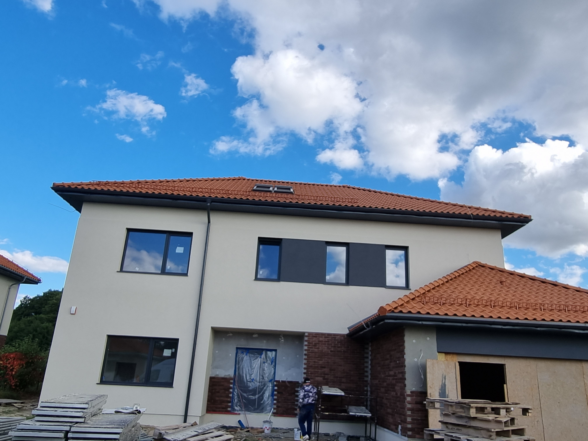 Dwukondygnacyjny dom z jasną elewacją i czerwoną dachówką w trakcie prac wykończeniowych. Widoczne materiały budowlane i pracownik przy elewacji z cegły.