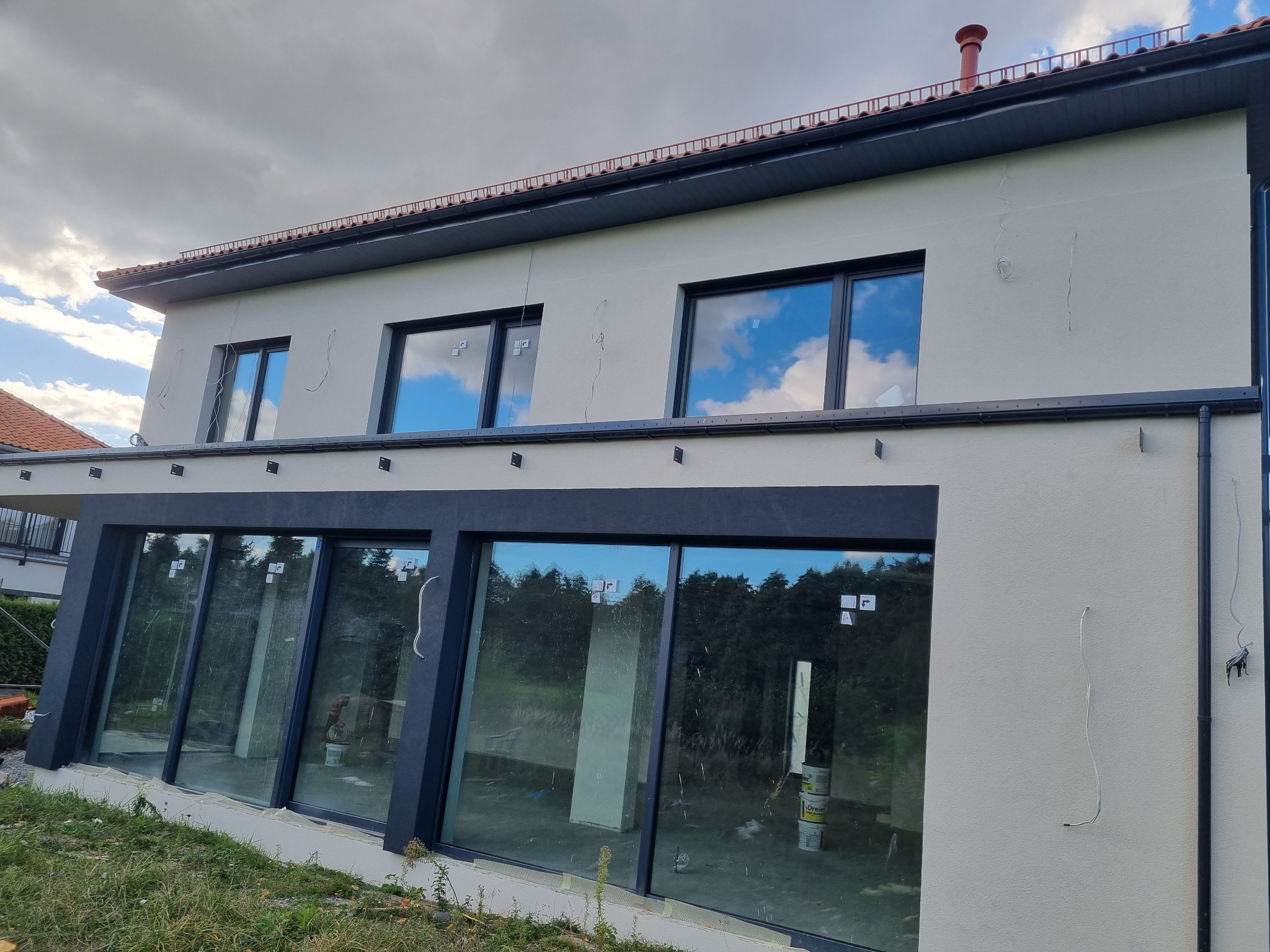 Elewacja domu jednorodzinnego z dużymi oknami, świeżo po malowaniu. Widoczne niedociągnięcia w postaci wystających kabli i śladów po szpachlowaniu. W oknach odbija się błękitne niebo.