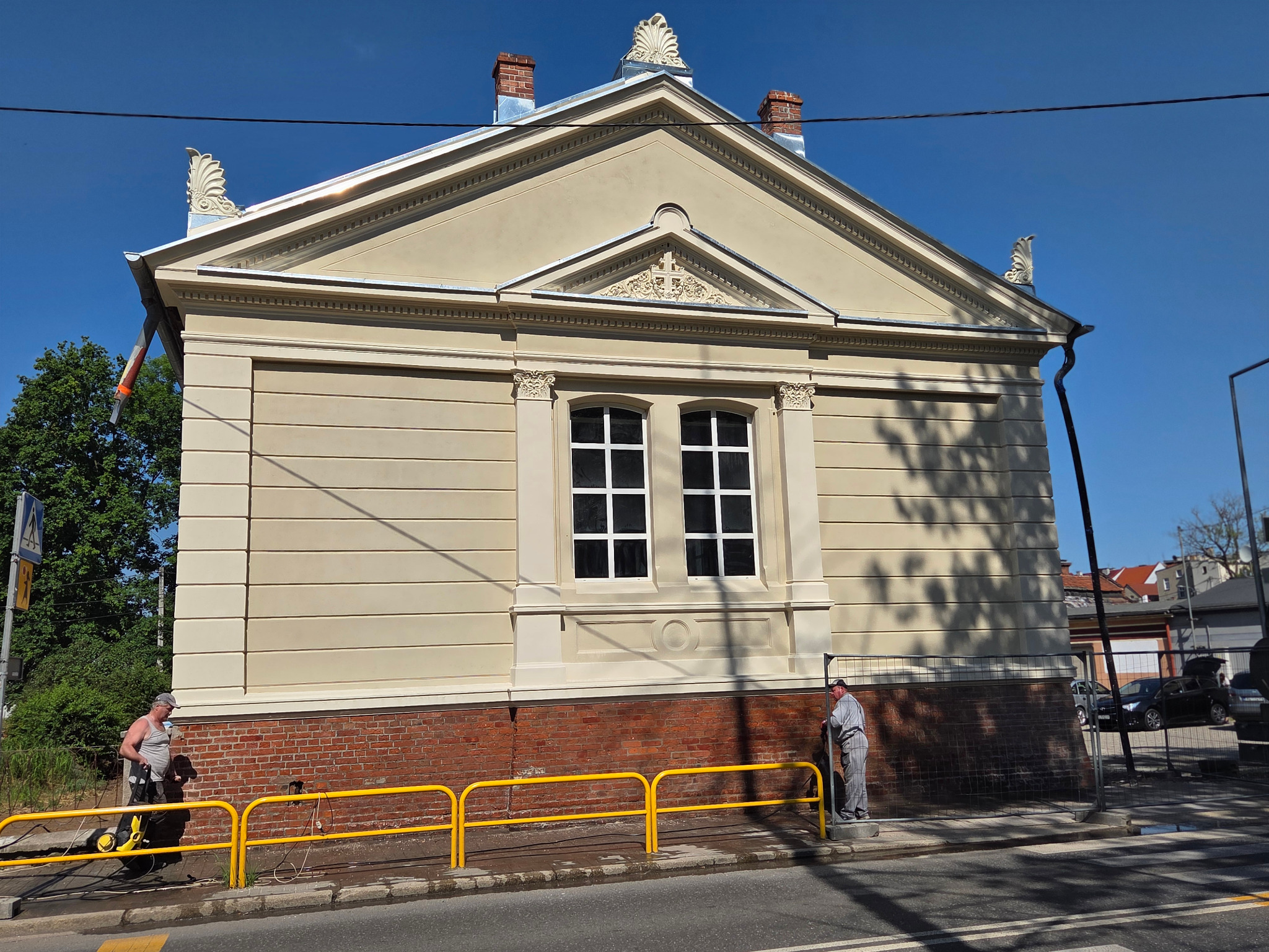 Elewacja budynku po renowacji, z detalami architektonicznymi, czyszczona myjką ciśnieniową. Widoczny fragment chodnika i żółte barierki zabezpieczające.