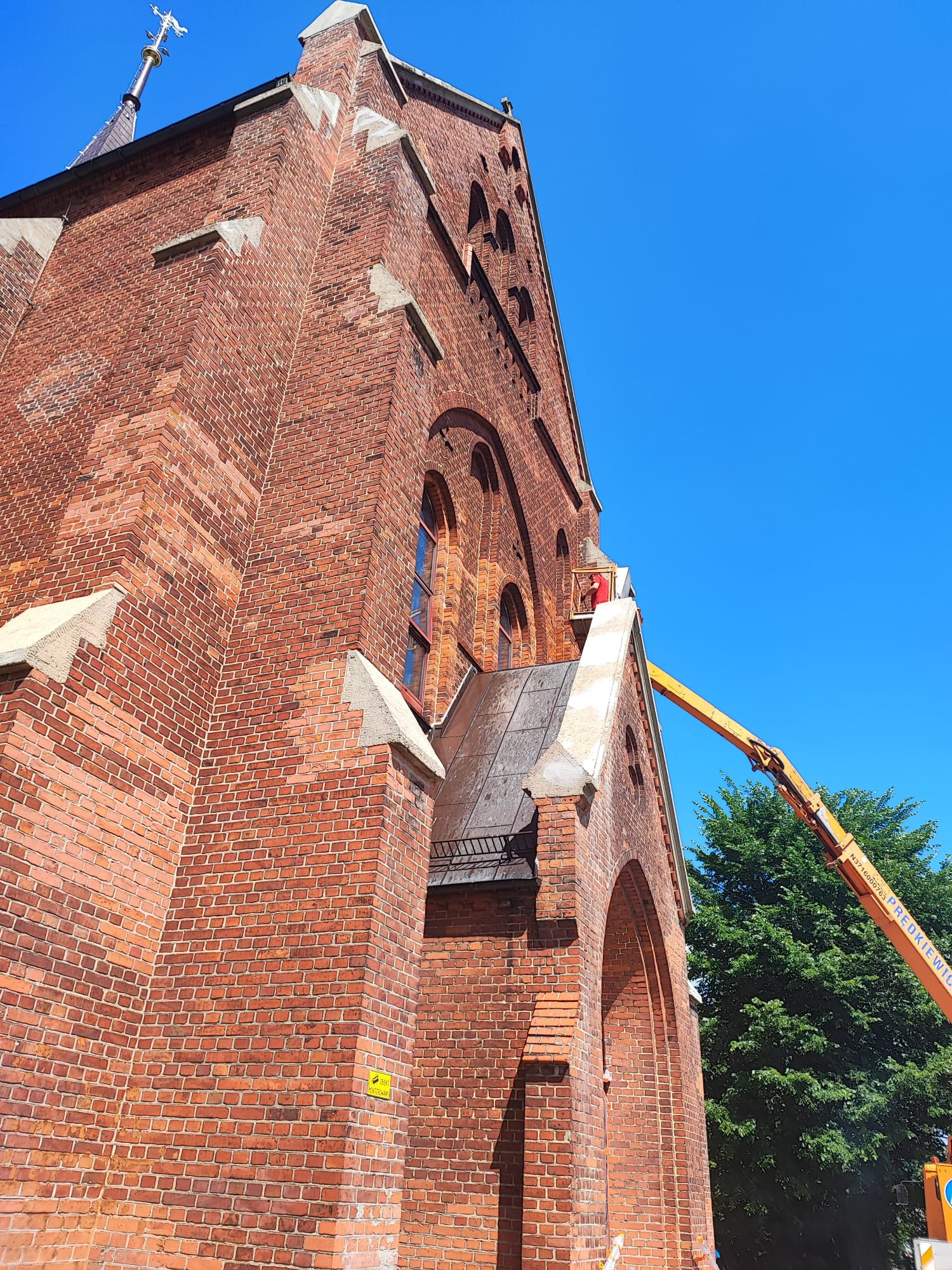Ceglana fasada kościoła w trakcie renowacji z użyciem podnośnika koszowego na tle błękitnego nieba i zieleni drzew. Widoczny fragment wieży i detale architektoniczne.