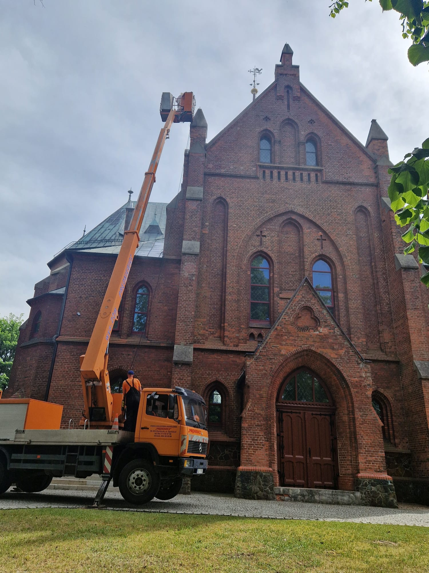 Prace na wysokościach przy ceglanej fasadzie kościoła z użyciem podnośnika koszowego. Widoczny fragment zieleni i błękitne niebo.