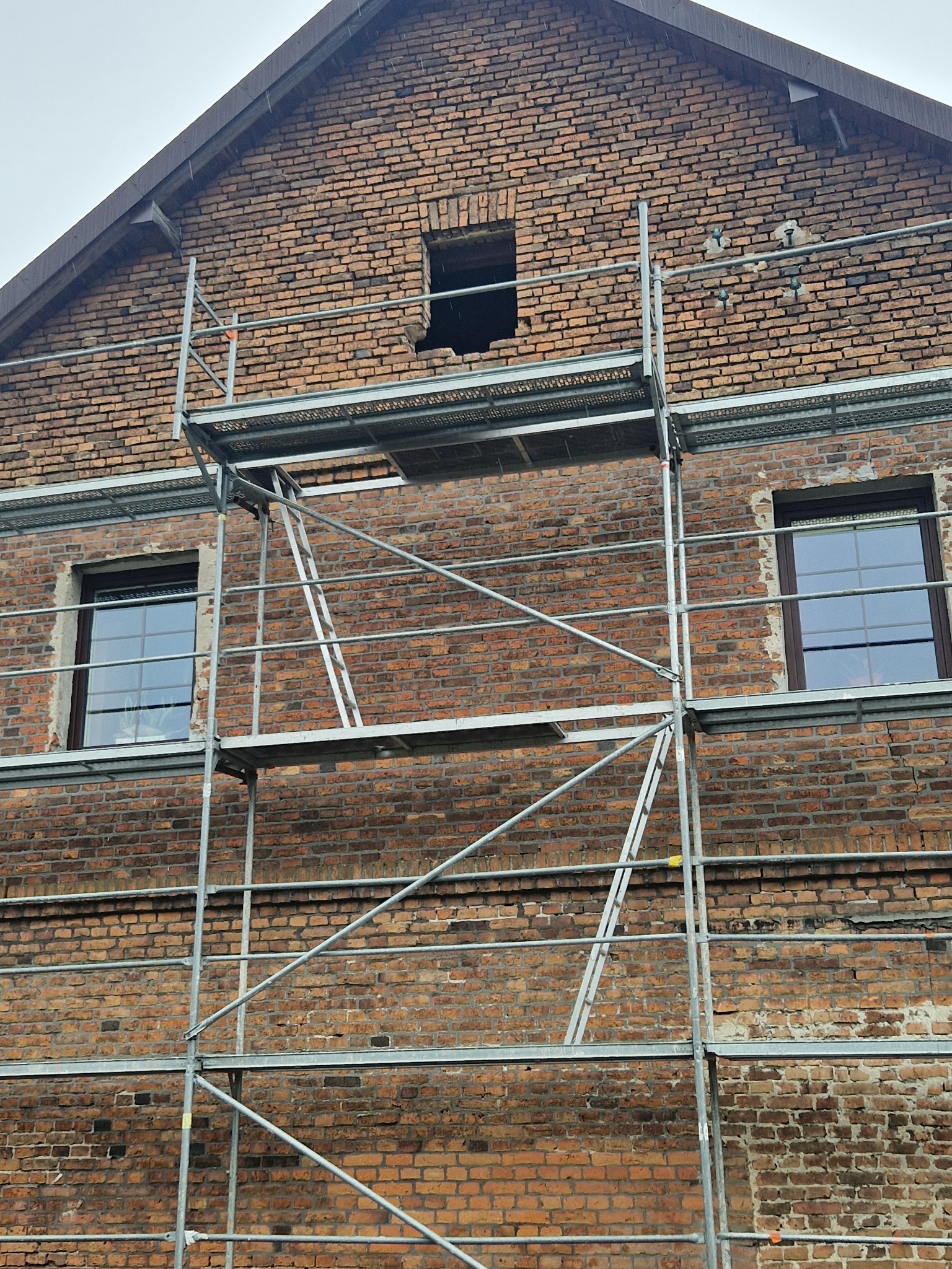 Ceglana elewacja budynku w trakcie renowacji, z widocznym rusztowaniem i oknami. Fragment ściany z uszkodzonym murem nad oknem. Prace remontowe w toku.