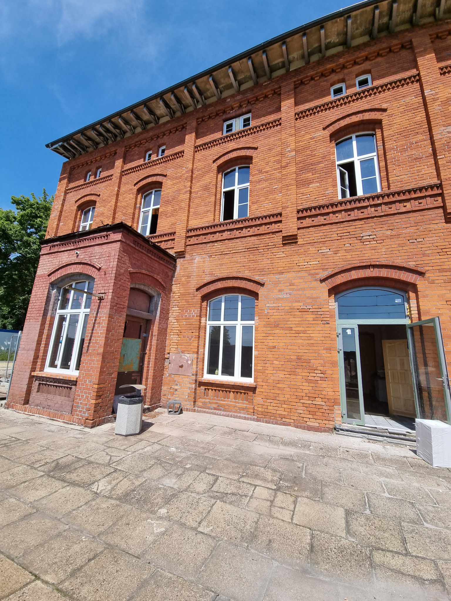 Ceglany budynek z białymi oknami i otwartymi drzwiami wejściowymi, widok z dołu, błękitne niebo w tle. Widoczny fragment chodnika z kamiennej kostki.