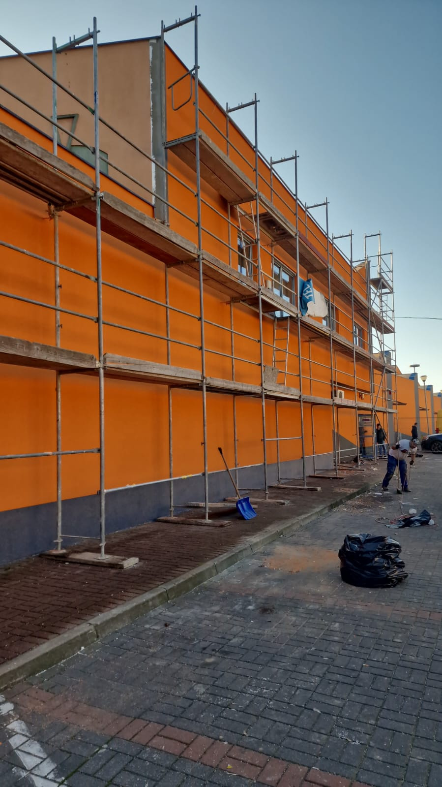 Pomarańczowa elewacja budynku w trakcie prac, widoczne rusztowania i pracownik. Na chodniku leży niebieska łopata i czarny worek na śmieci.