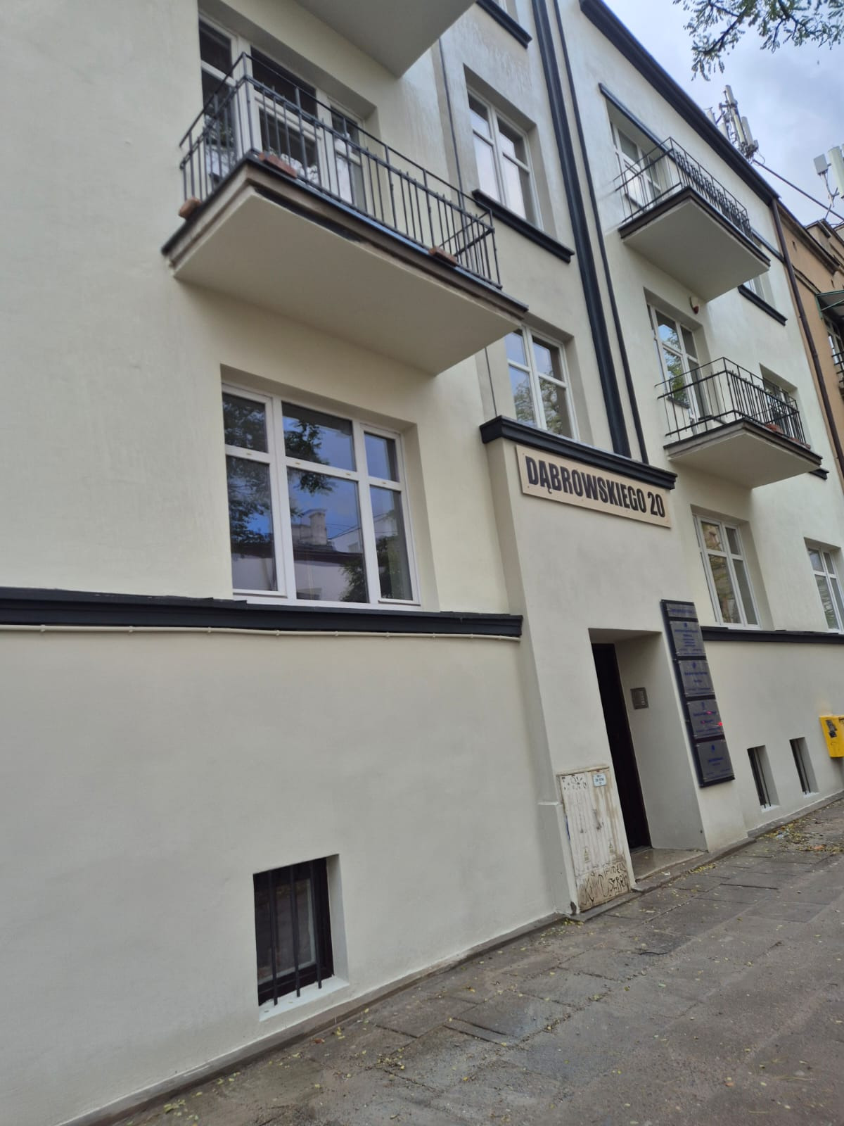 Odnowiona elewacja kamienicy z balkonami i tabliczką adresową 'Dąbrowskiego 20', po malowaniu na jasny kolor z czarnymi akcentami.