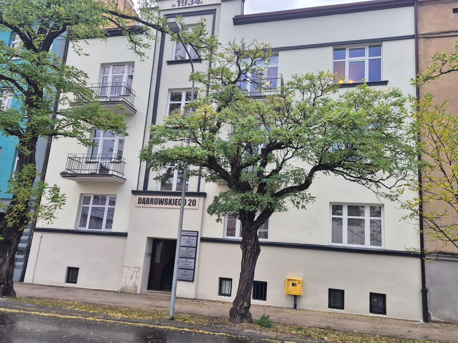 Odnowiona fasada kamienicy z 1934 roku, z jasną elewacją i ciemnymi akcentami, widoczna spod rozłożystego drzewa. Tabliczka z adresem 'Dąbrowskiego 20'.