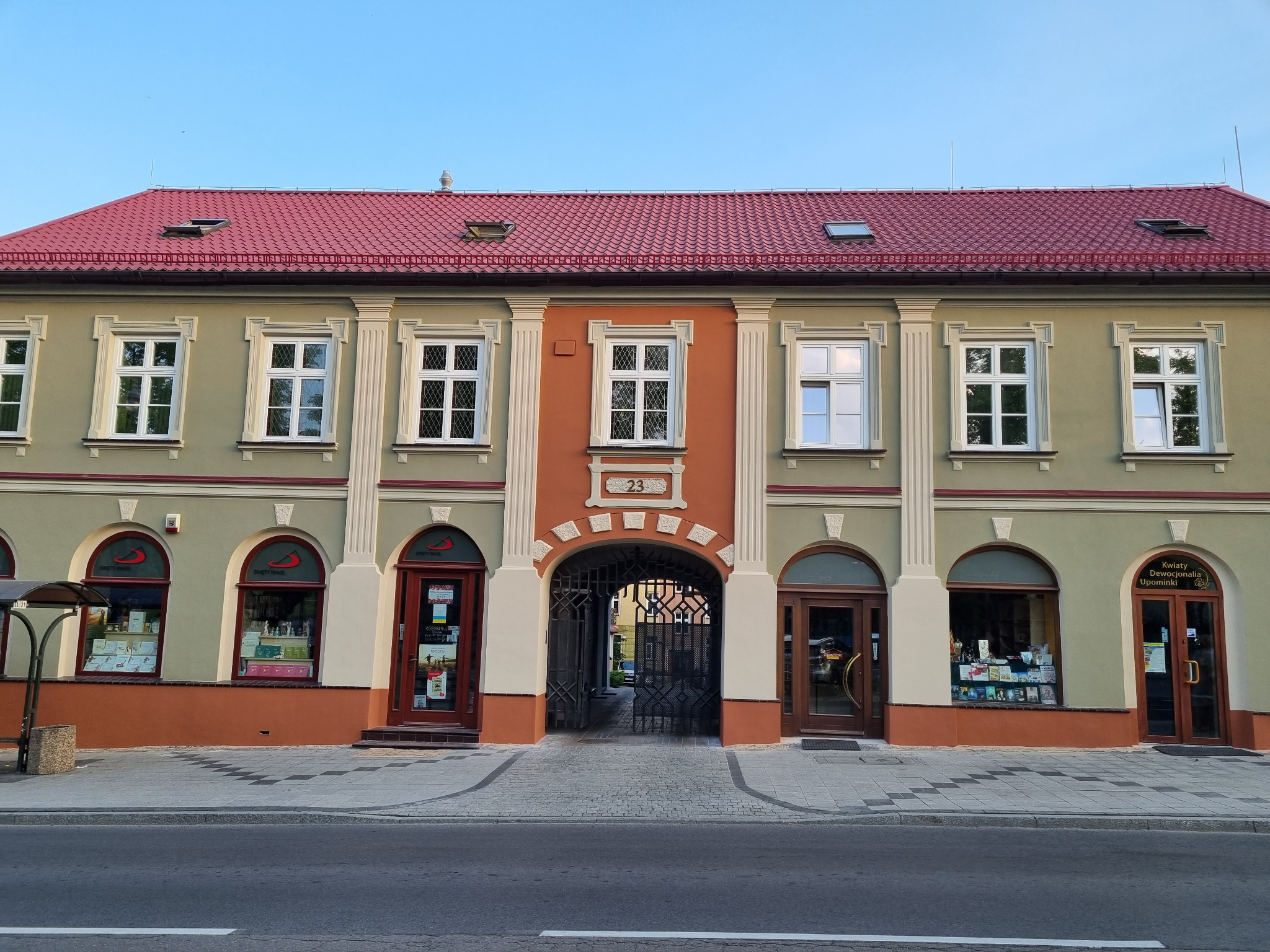 Odnowiona fasada kamienicy z czerwonym dachem, widoczne okna z białymi ramami i dekoracyjne elementy architektoniczne. Uwagę przykuwa numer 23 nad bramą.