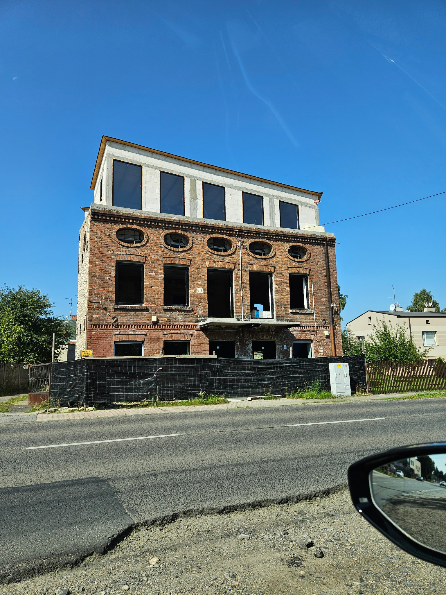 Dwupiętrowy budynek z czerwonej cegły w trakcie remontu, z nową nadbudową z białych pustaków i dużymi oknami, otoczony czarną siatką. Widok z ulicy, błękitne niebo.
