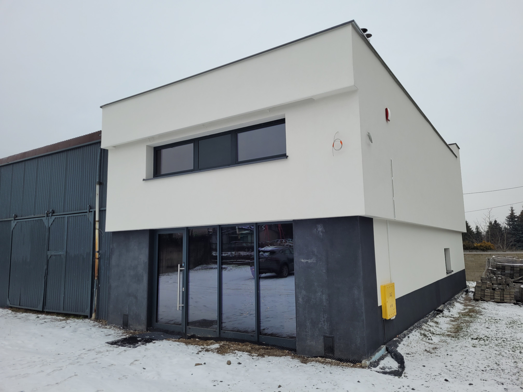 Dwukondygnacyjny budynek z białą elewacją i ciemnoszarym cokołem, duże okna, zimowy krajobraz. Widoczne elementy instalacji na elewacji.