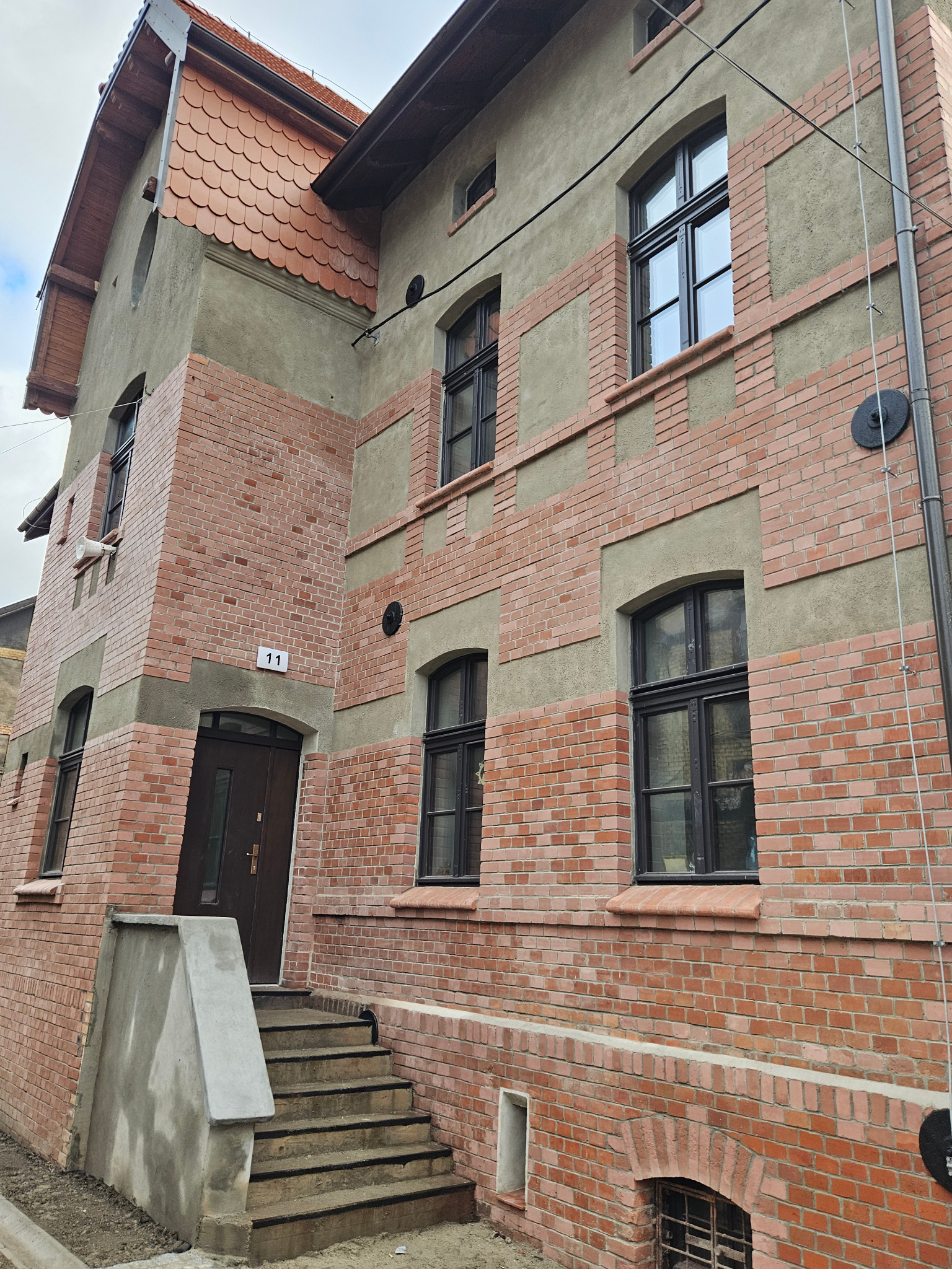 Odnowiona elewacja kamienicy z ceglanymi elementami i szarym tynkiem. Widoczne okna z czarnymi ramami i numer budynku '11'.