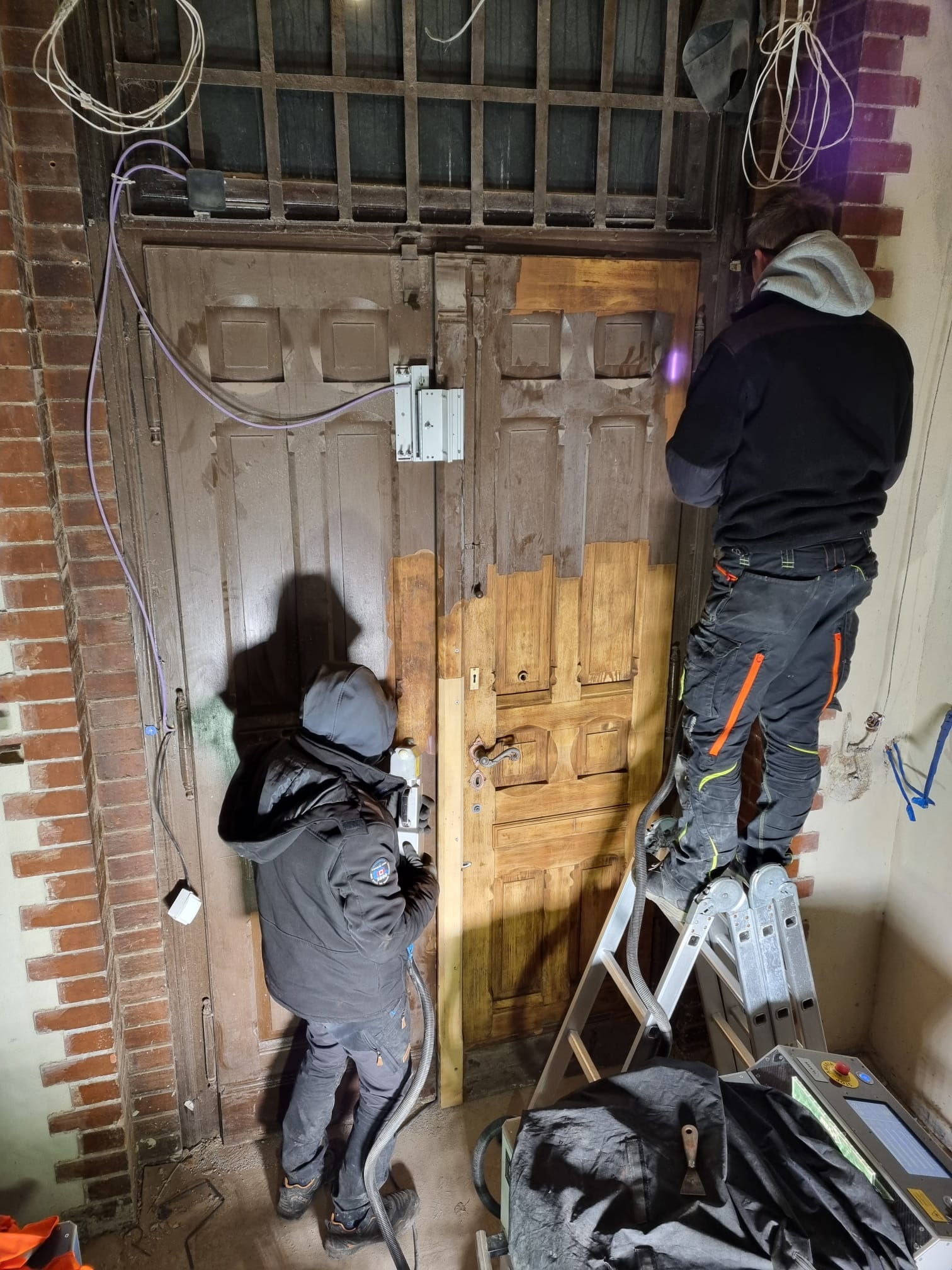 Renowacja drewnianych drzwi wejściowych metodą laserową. Dwóch pracowników usuwa starą farbę, stojąc na drabinie. Widoczny efekt czyszczenia laserem.