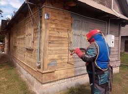 Pracownik w kombinezonie ochronnym czyści drewnianą elewację domu metodą strumieniowo-ścierną, przygotowując ją do malowania. Widoczny fragment elewacji po czyszczeniu.