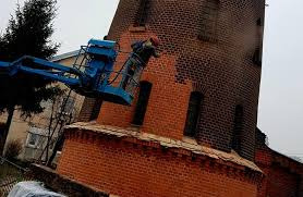 Renowacja ceglanej wieży: malarz na podnośniku koszowym, odnawia elewację, widoczne fragmenty pomalowanej i surowej cegły, pochmurne niebo w tle.
