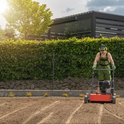Botanik Karolina Kwaśnik - Pracownik w zielonym kombinezonie aeruje glebę aeratorem na przygotowanym terenie pod trawnik, w tle żywopłot i nowoczesna pergola.