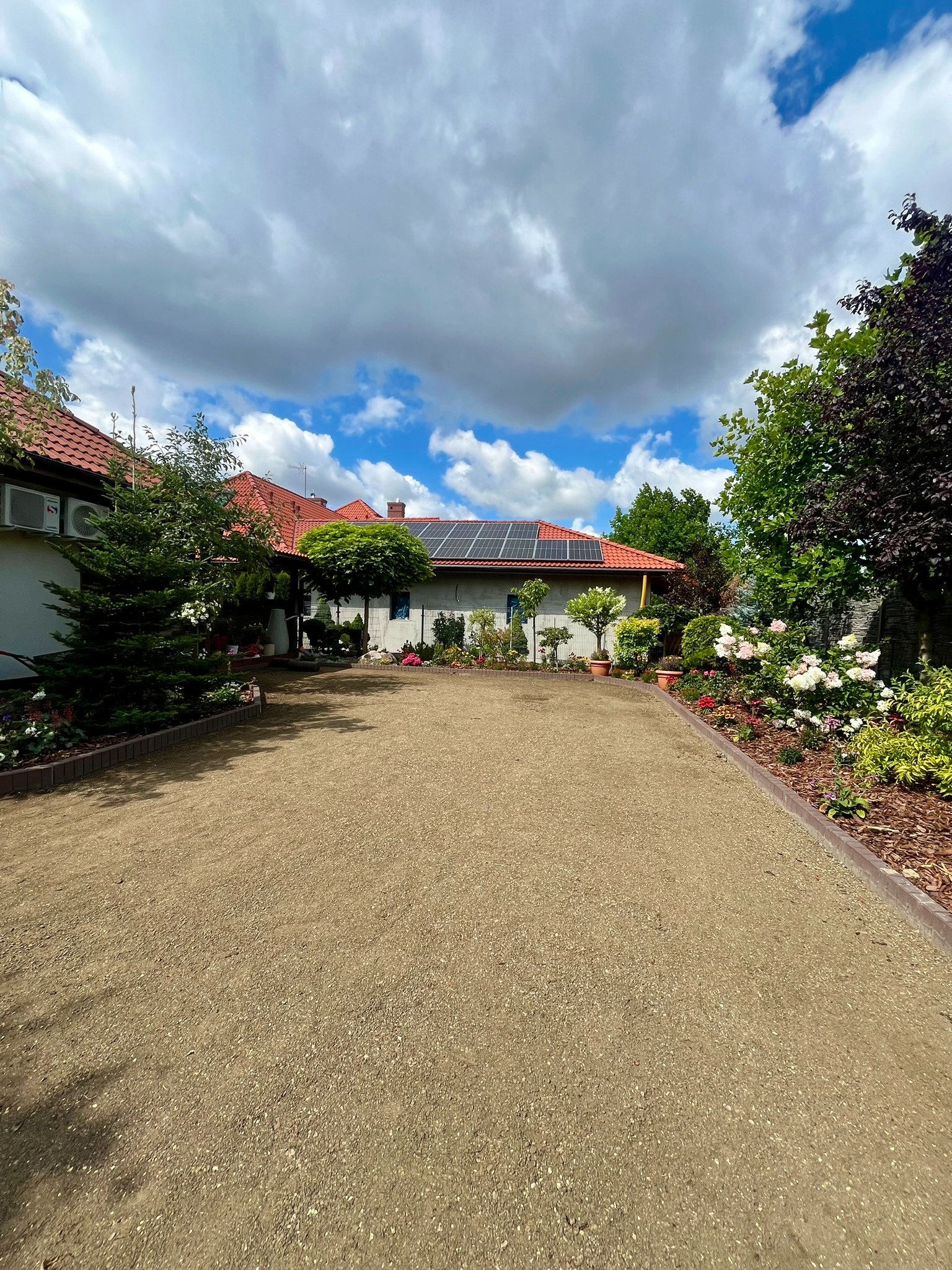 Ukończony podjazd z drobnym kruszywem, obrzeża z kostki, zadbany ogród z kwitnącymi różami i drzewami, dom z panelami słonecznymi na dachu w tle.