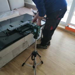 Jarek Król - Instalator w trakcie montażu uchwytu ściennego do telewizora, używając poziomicy laserowej na statywie. Widoczny telewizor leżący na kanapie i elementy montażowe.