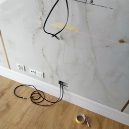 Jarek Król - Instalacja kabli w ścianie wykończonej płytkami imitującymi marmur. Widoczne gniazdka, listwa przypodłogowa, podłoga z paneli i narzędzia: taśma malarska i ołówek.