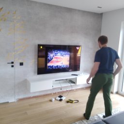 Jarek Król - Telewizor na ścianie z podłączonymi kablami i urządzeniami. Widoczny mężczyzna w tle. Instalacja sprzętu RTV w nowoczesnym wnętrzu.