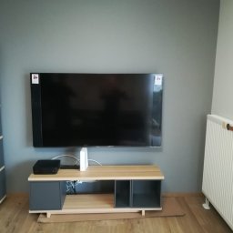 Jarek Król - Telewizor na ścianie nad szafką RTV, obok regał i grzejnik. Widoczne kable i urządzenia podłączone do TV. Szaro-beżowa kolorystyka wnętrza.