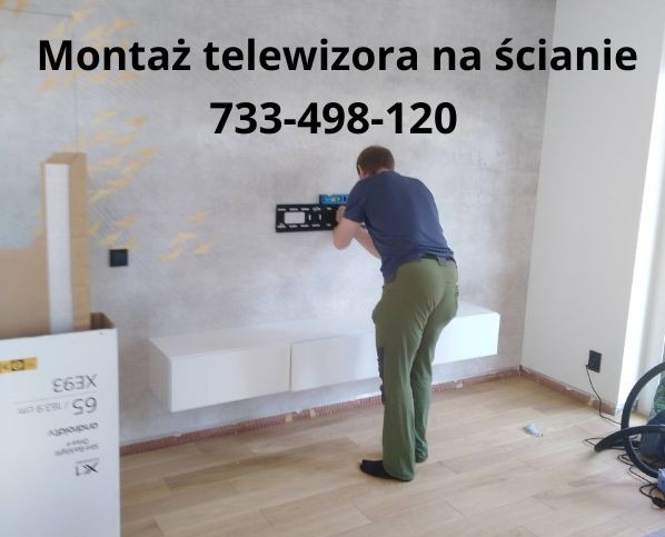 Instalator montuje uchwyt TV na ścianie w nowoczesnym salonie. Widoczna biała szafka RTV i karton po telewizorze. Na ścianie numer telefonu.